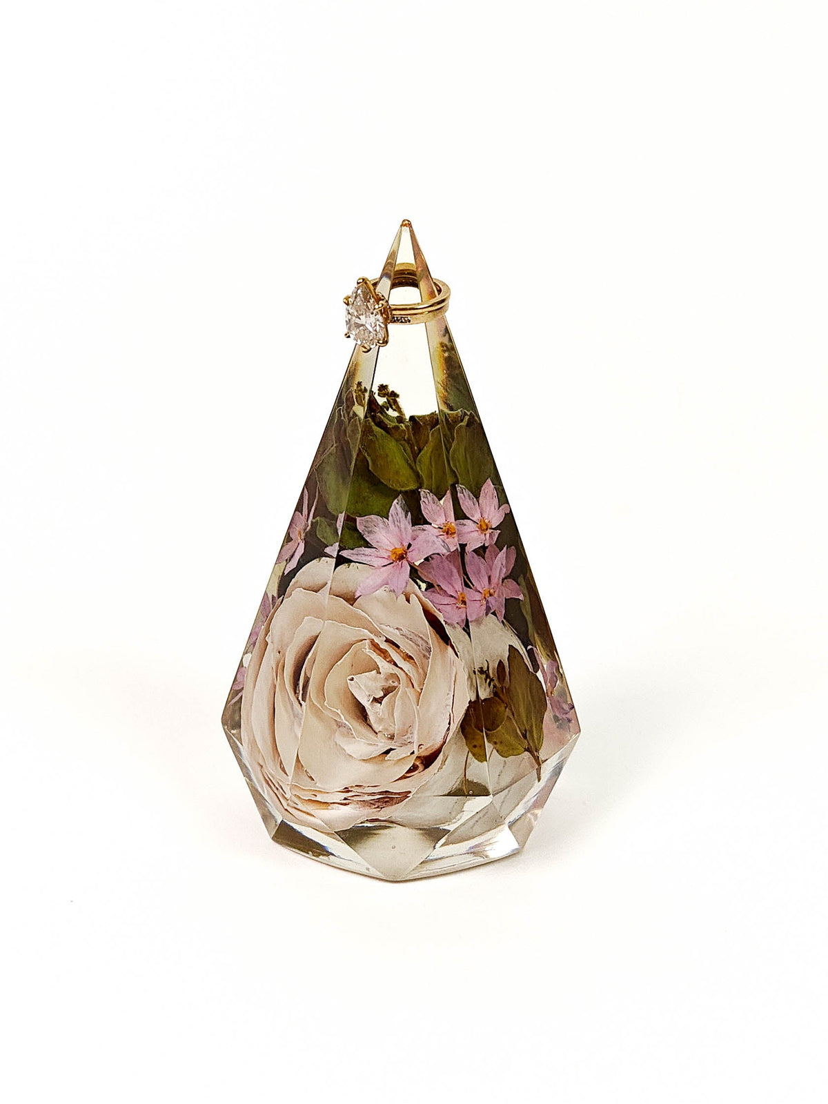 A faceted clear resin keepsake urn or pendant with dried flowers, featuring a large cream rose and pink blossoms inside a gold-toned cap, set against a plain white background.