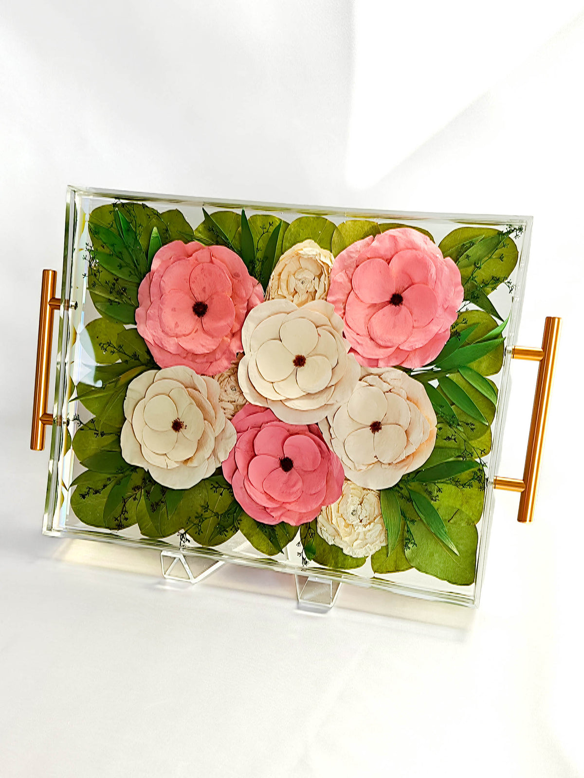 A rectangular clear acrylic decorative tray with gold side handles, filled with artificial pink and cream flowers and green leaves, displayed on a white background.