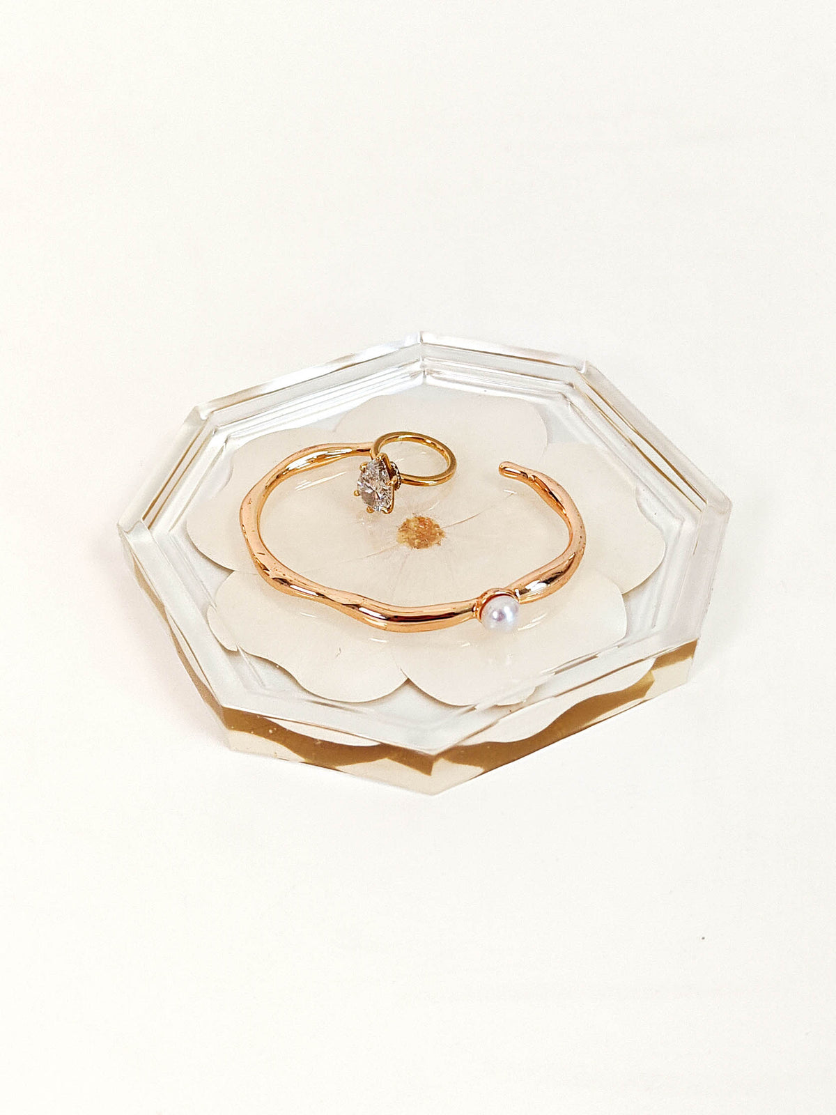 A rose gold open cuff bracelet with a small clear gemstone and a white pearl, displayed on a scalloped clear acrylic tray with a gold base, set against a plain white background.