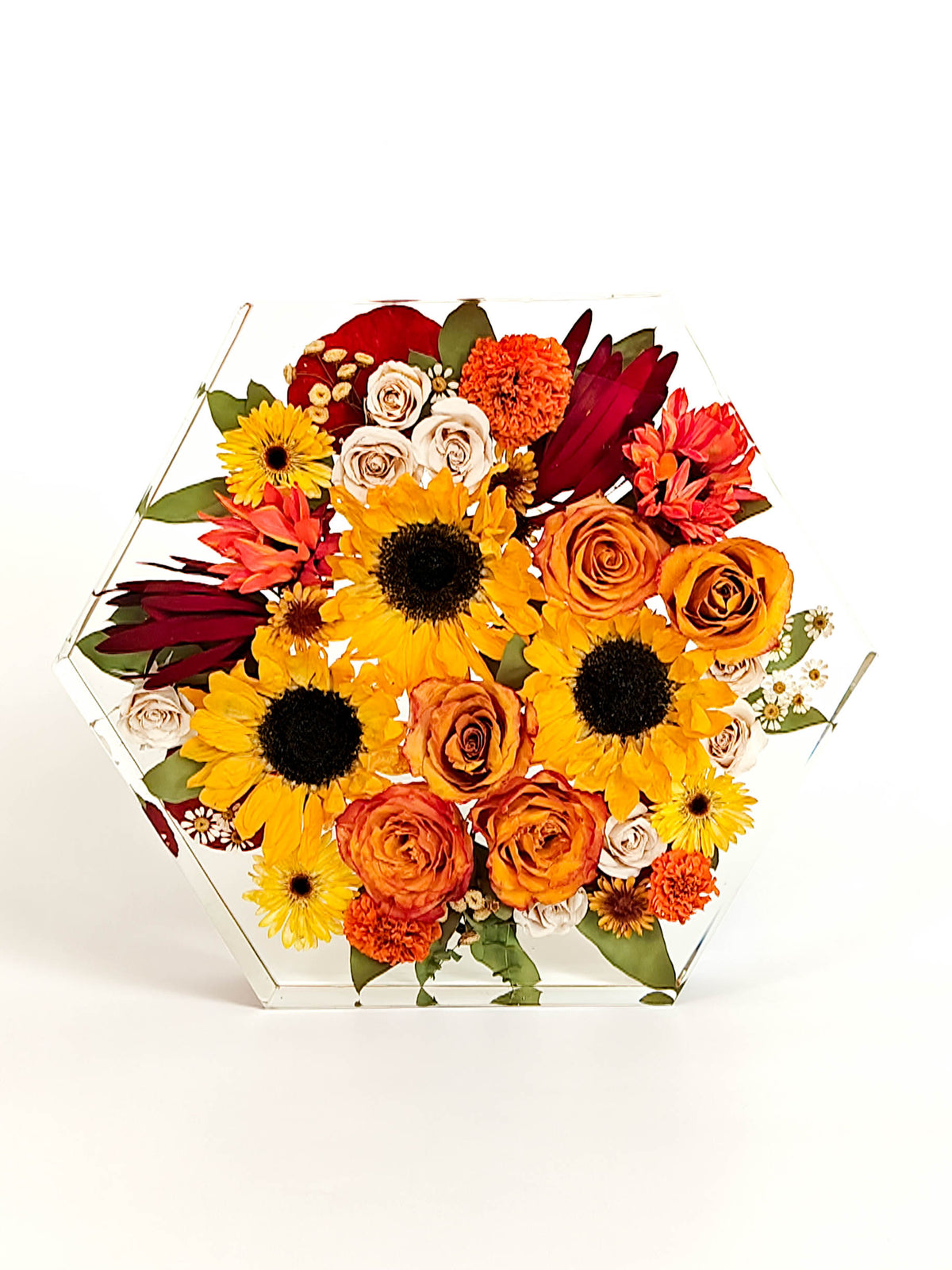 A vibrant artificial flower arrangement in a clear hexagonal acrylic container, featuring yellow sunflowers, orange and red roses, marigolds, and burgundy foliage, set against a plain white background.