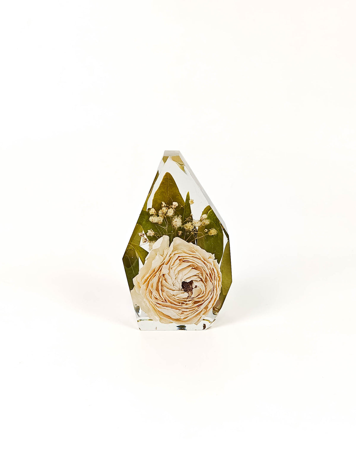 A faceted clear resin crystal vase containing a dried cream-colored rose, green leaves, and small white flowers, set against a plain white background.