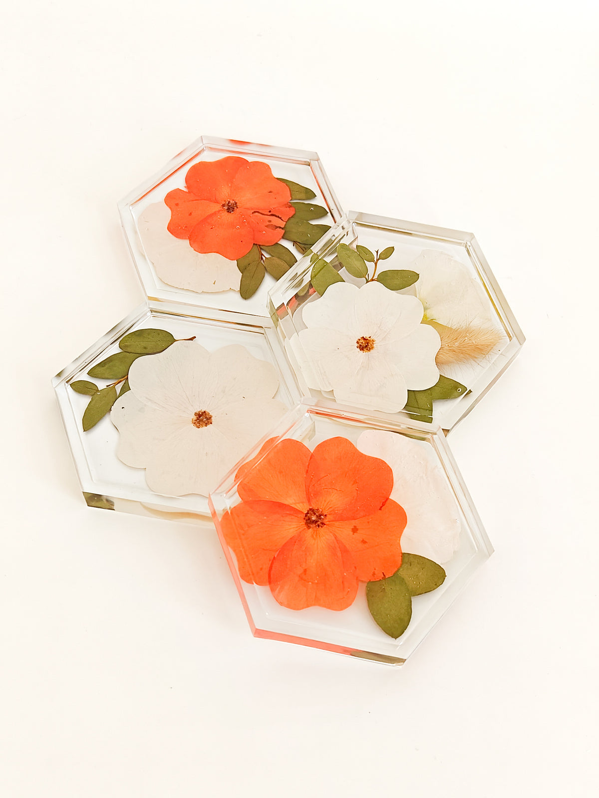 Four clear hexagonal resin coasters with pressed flowers, including two orange flowers and two white flowers with green leaves, arranged on a white background.