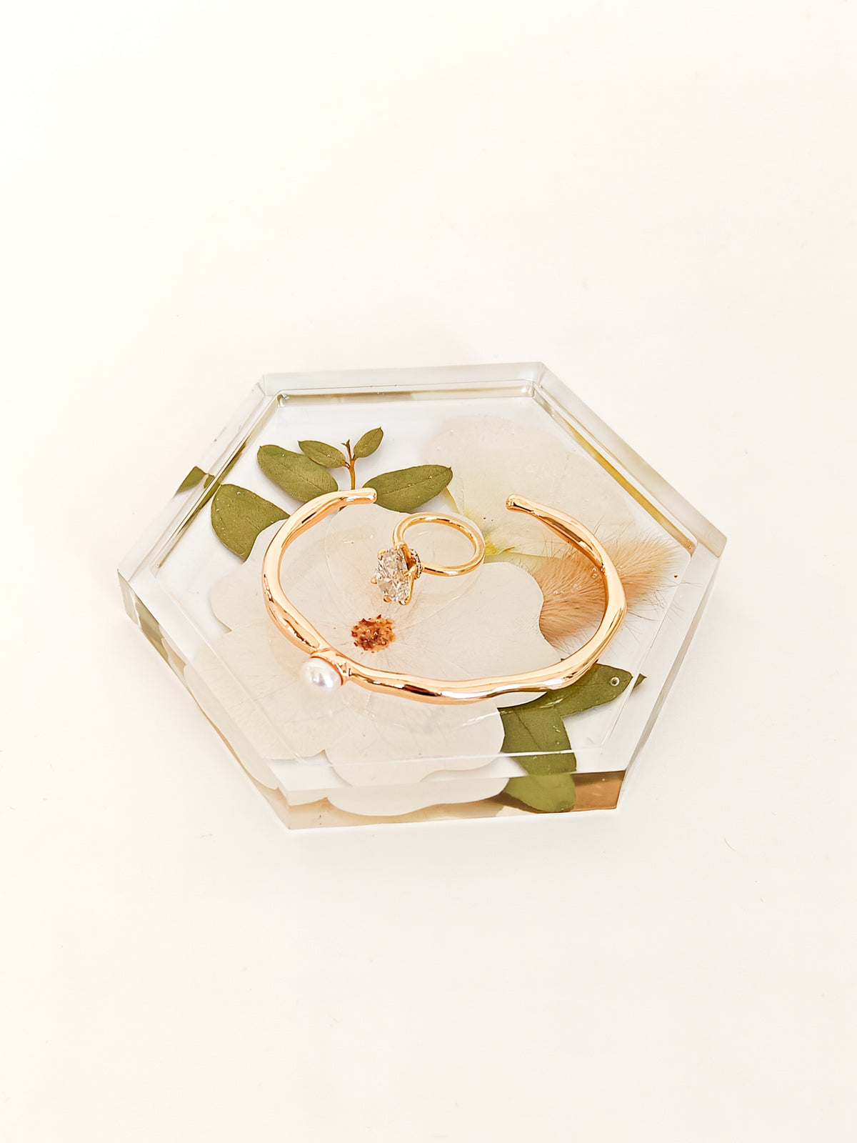 A hexagonal clear resin tray with dried green leaves and white flowers, holding a gold cuff bracelet and two gold rings, photographed on a clean white background.