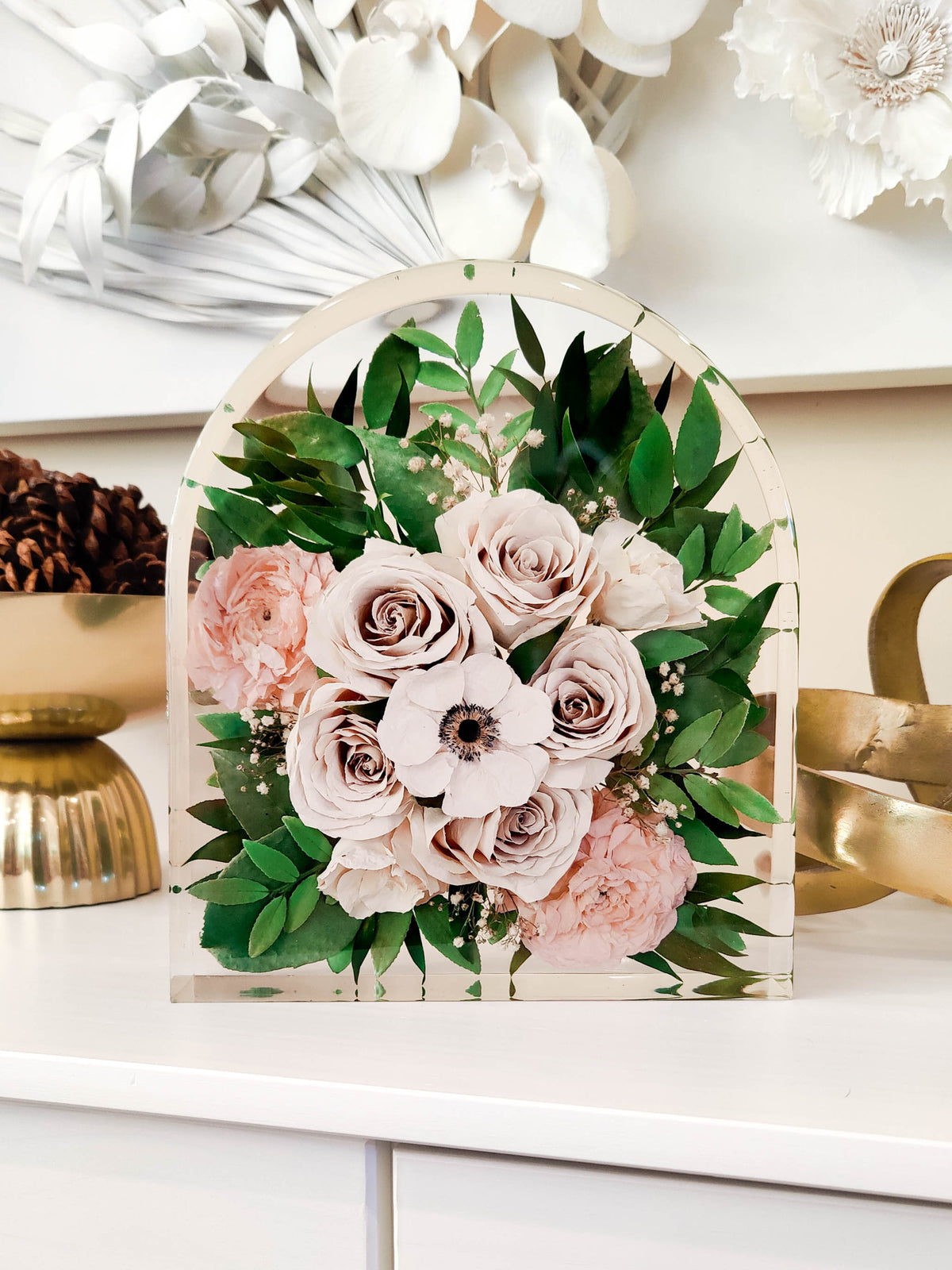 A decorative acrylic arch plaque featuring a preserved floral arrangement with blush pink roses, green foliage, and white anemone flowers, displayed on a white shelf.
