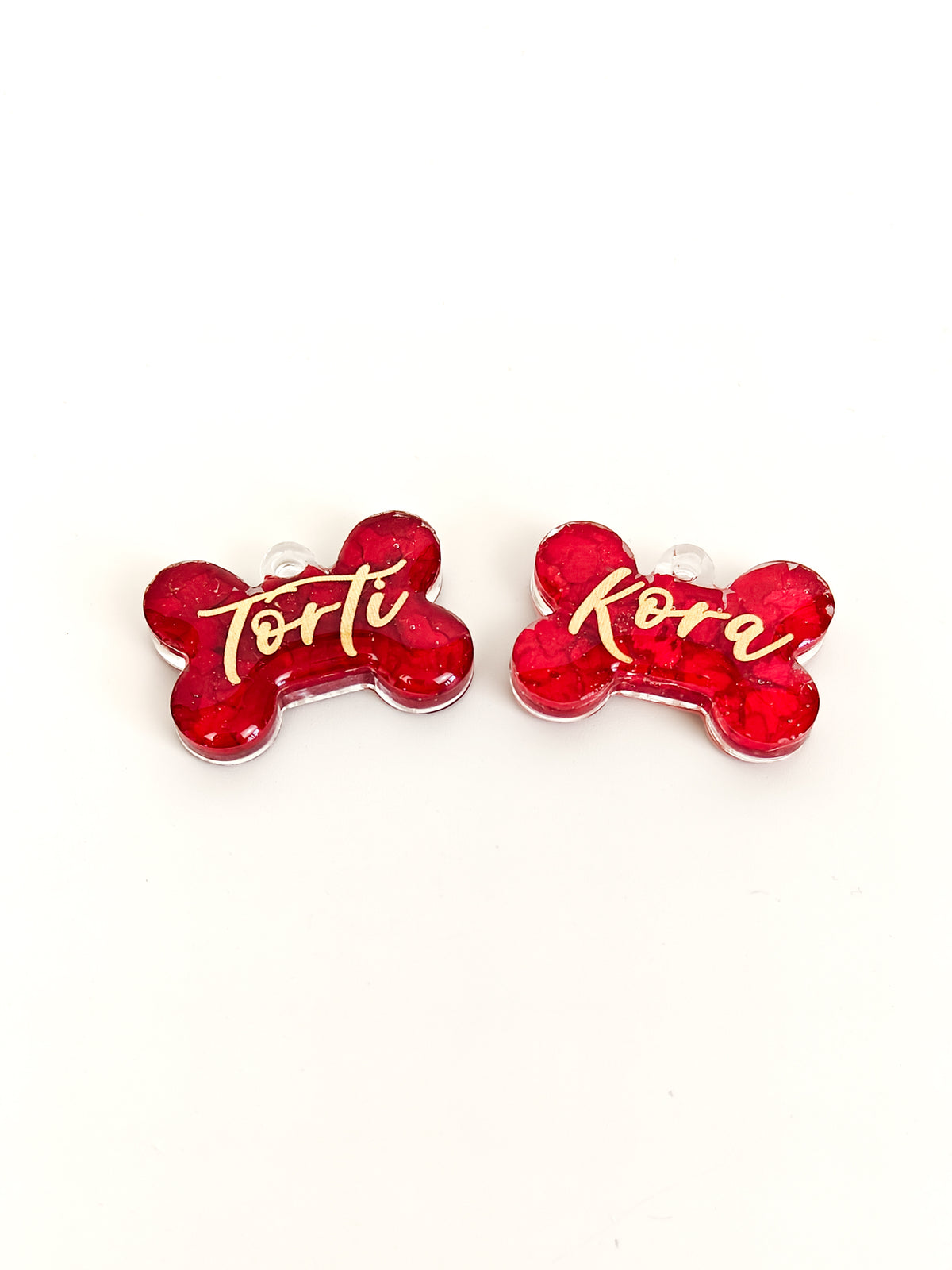 Two red bone-shaped pet nameplates with glittery red fill and gold script lettering, displayed on a clean white background. One is labeled 'Torti' and the other 'Kora'.
