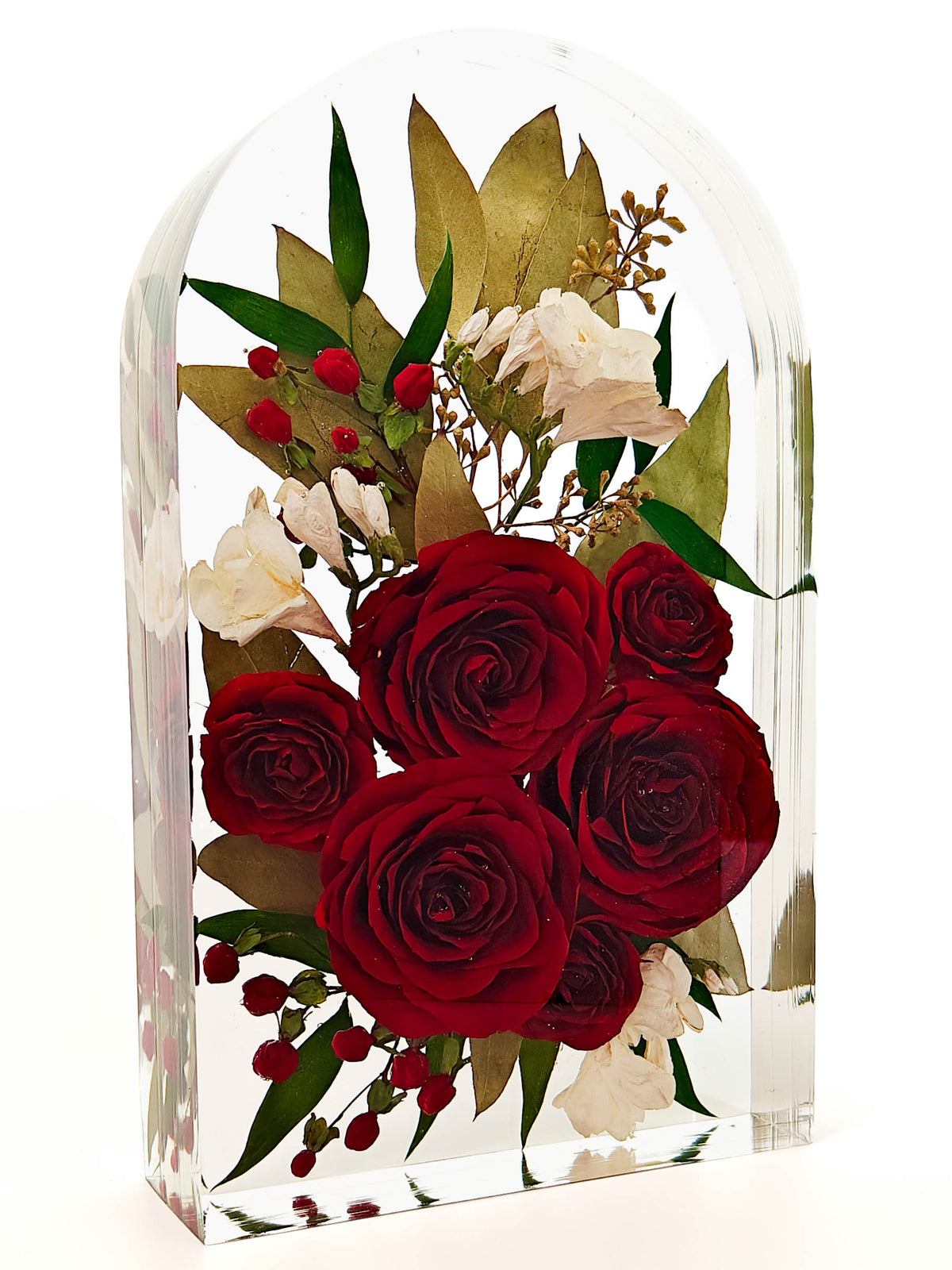 A clear acrylic arch-shaped flower holder containing a preserved floral arrangement with deep red roses, white blossoms, green leaves, and small red berries, photographed on a white background.