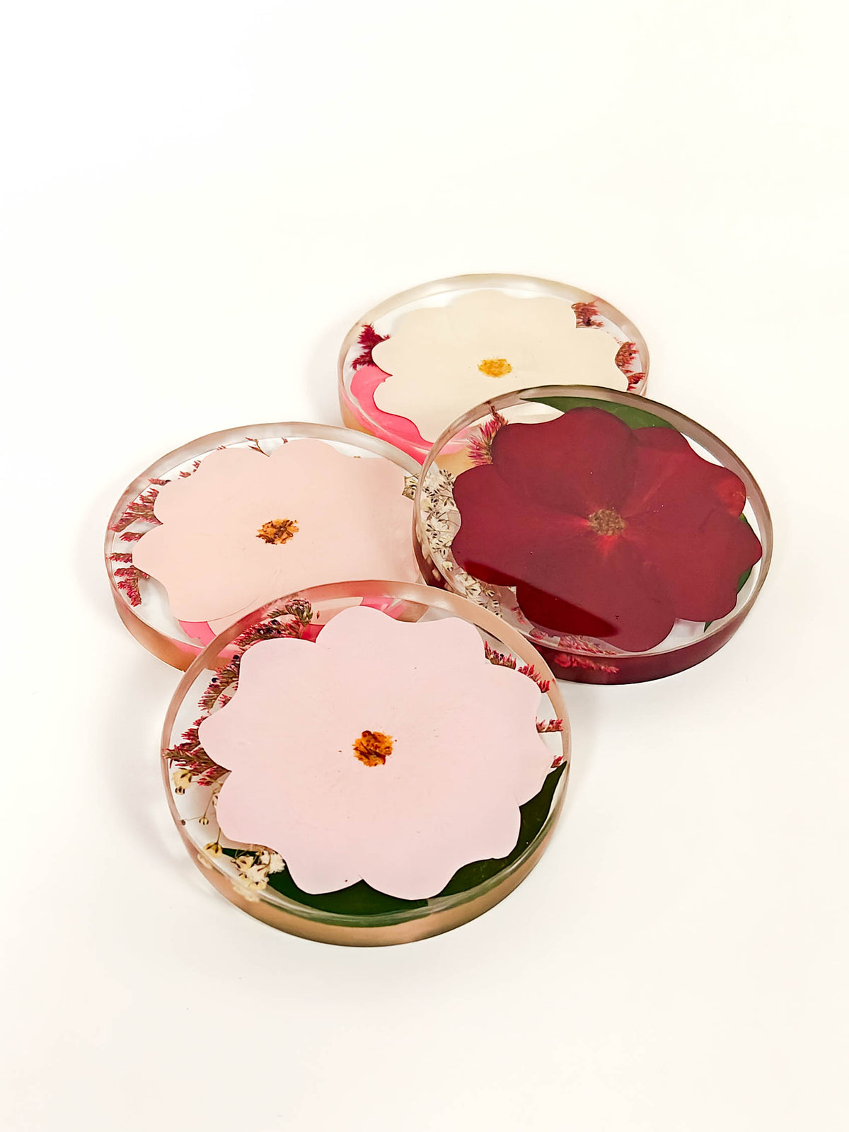 Four round resin coasters with embedded pressed flowers, arranged on a clean white background. The coasters feature pink, red, and cream flowers with green leaves and small gold accents.