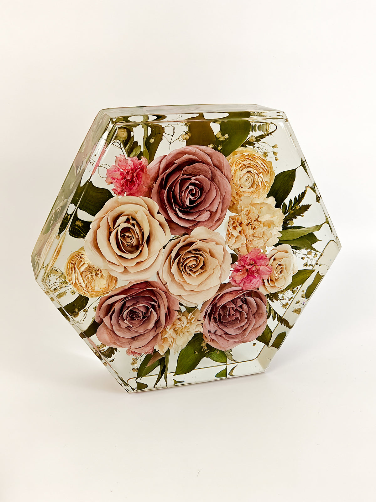 A hexagonal clear resin box containing a pressed floral arrangement with dusty pink, cream, and peach roses, green leaves, and small pink blossoms, photographed on a clean white background.
