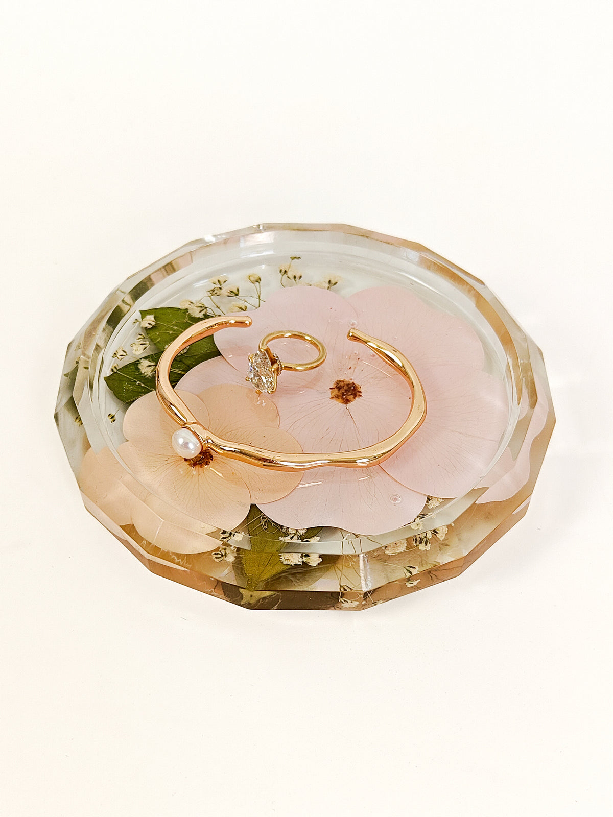 A gold-tone open cuff bracelet with a small pearl accent and tiny clear stones, displayed inside a faceted clear resin tray with pink flower petals and green leaves on a white background.