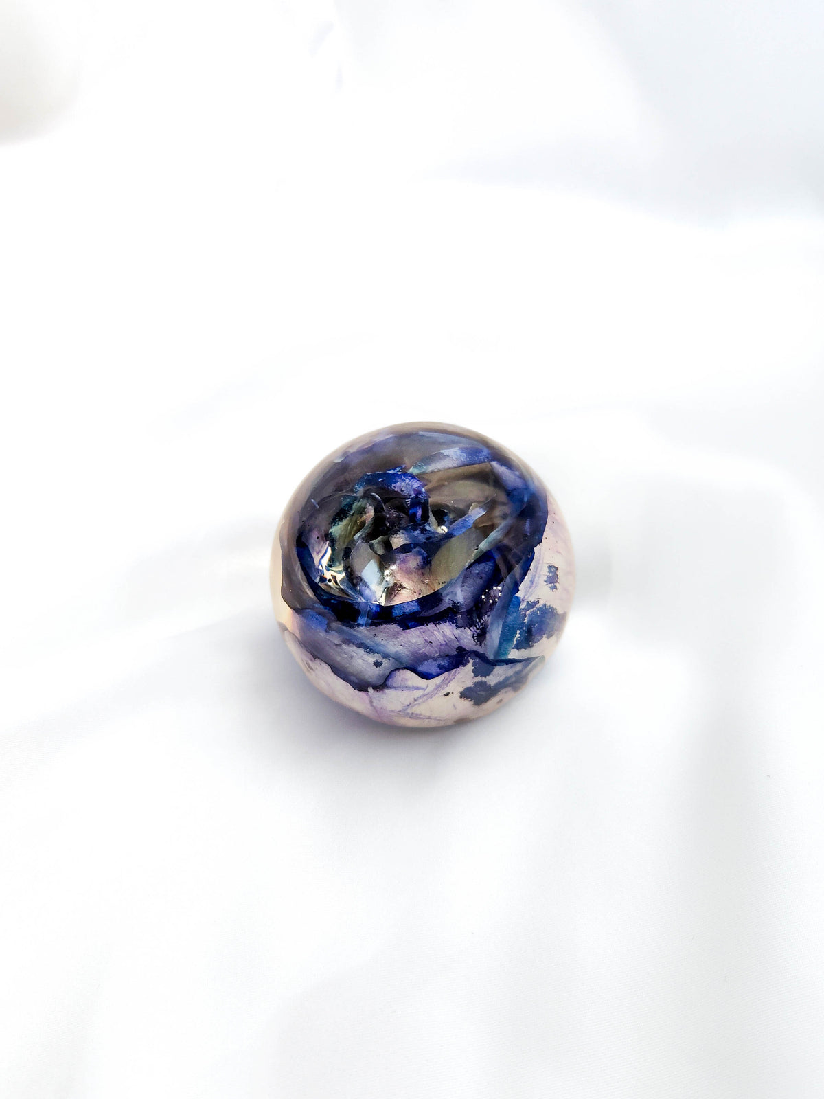 A single round glass bead with a marbled pattern of blue, purple, black, and white, resting on a white fabric background.