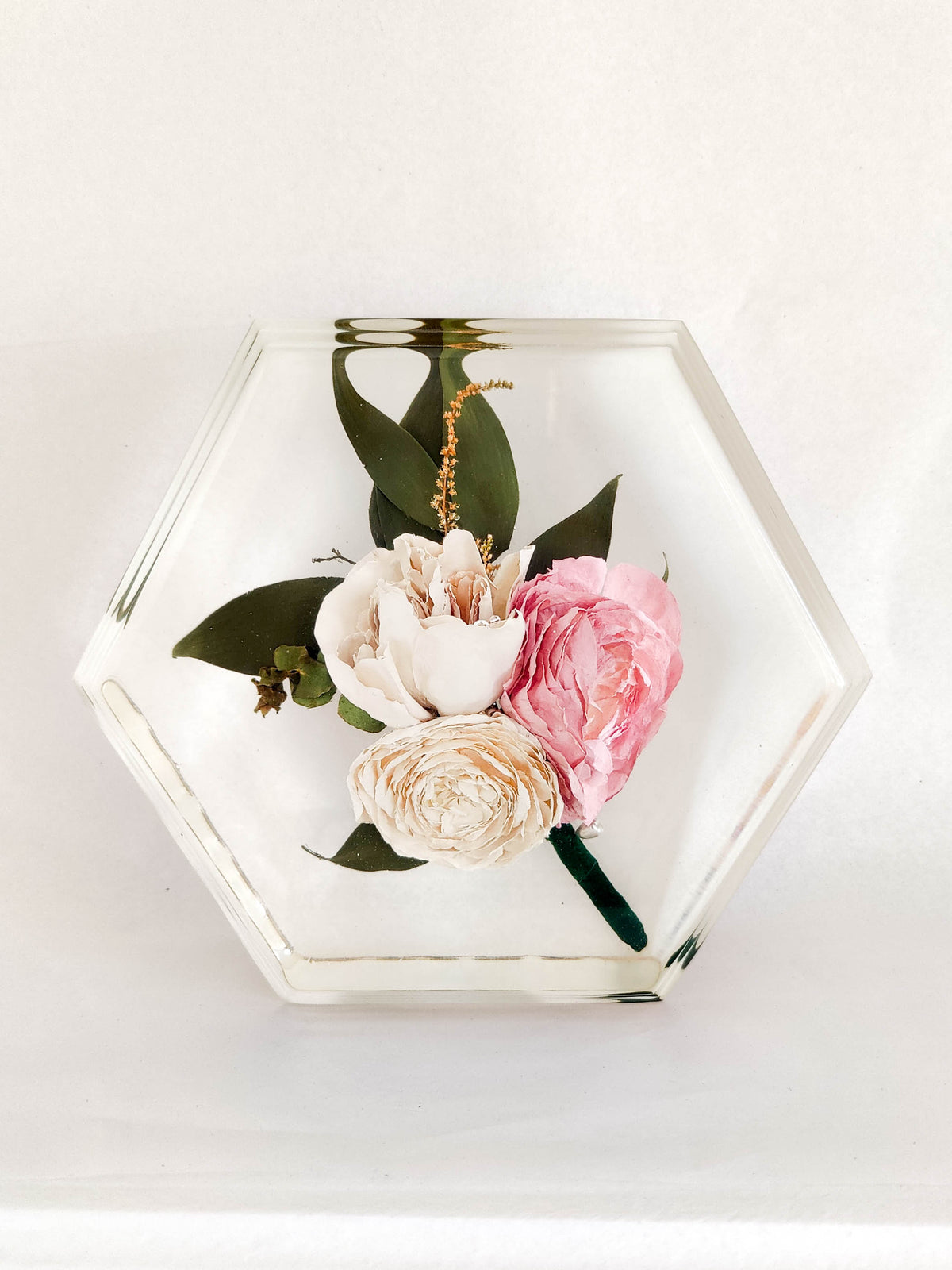 A hexagonal clear resin tray containing a pressed floral arrangement with pink and cream roses and green leaves, photographed on a white background.