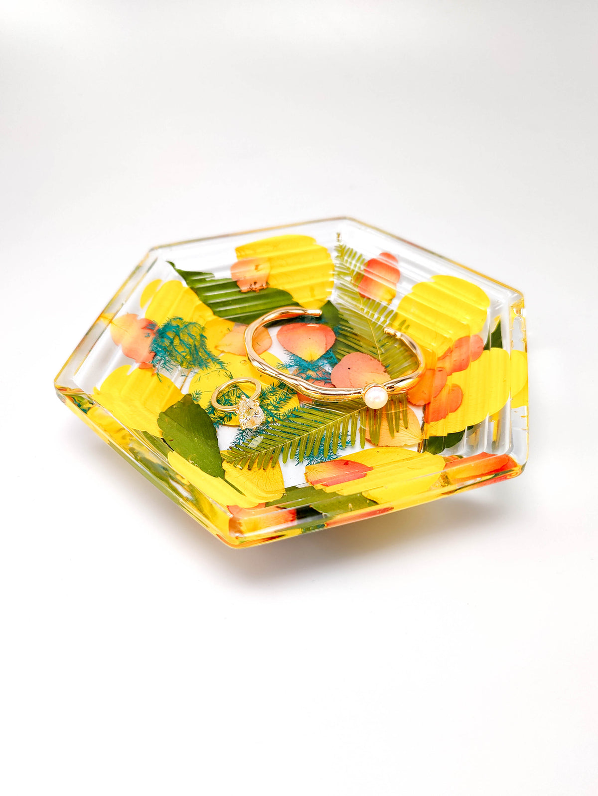 A clear hexagonal acrylic or glass dish containing pressed yellow flowers, green leaves, and a small orange flower, photographed on a clean white background.