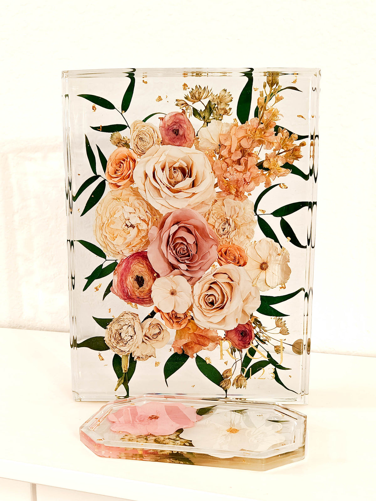 A clear acrylic plaque or tray featuring a pressed floral bouquet with peach, pink, cream, and gold accents, displayed on a white surface with a matching clear hexagonal base in front.