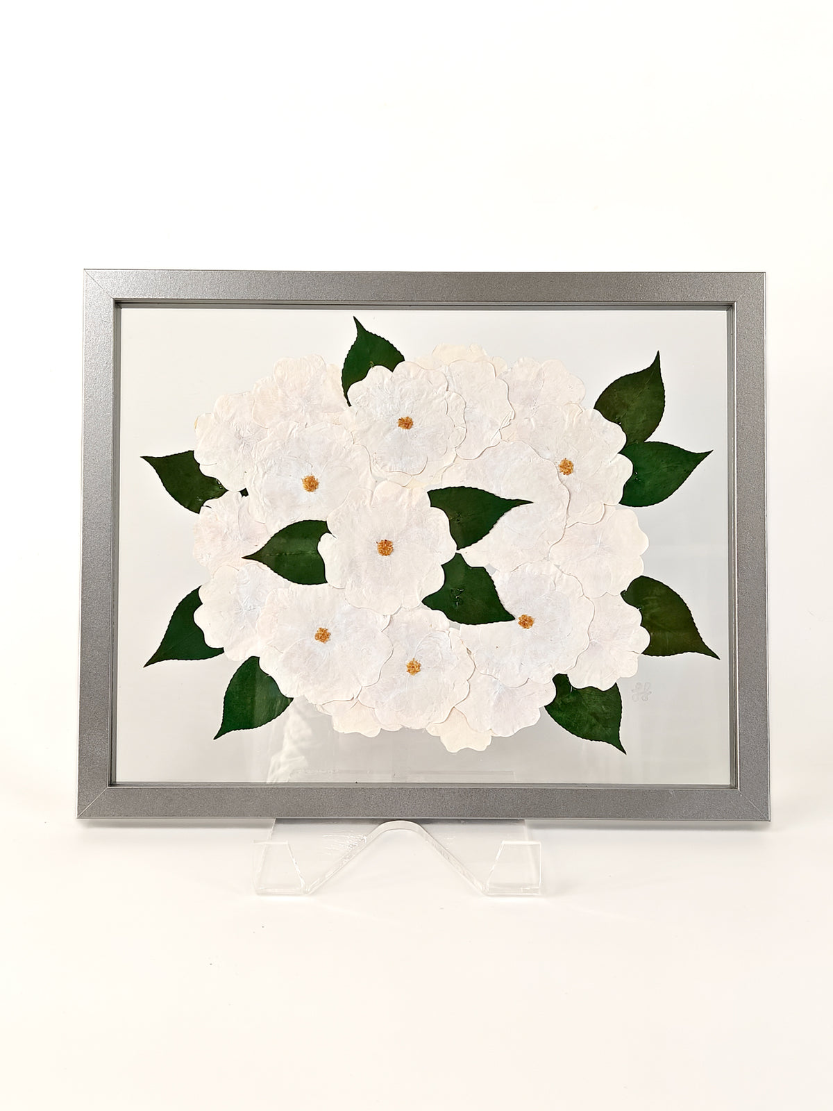 A framed floral artwork featuring layered white flower shapes with dark green leaves and small gold centers, displayed on a clear stand against a white background.