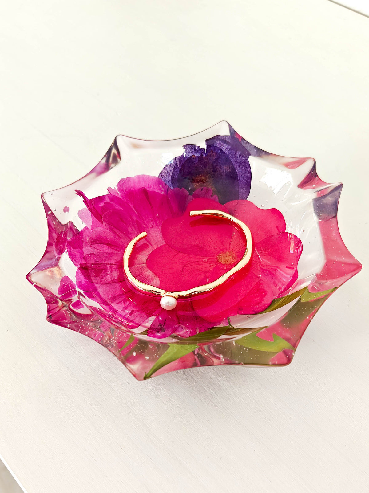 A gold bangle bracelet with a small white pearl bead rests inside a decorative, scalloped clear glass bowl filled with pressed pink and purple flowers on a white surface.