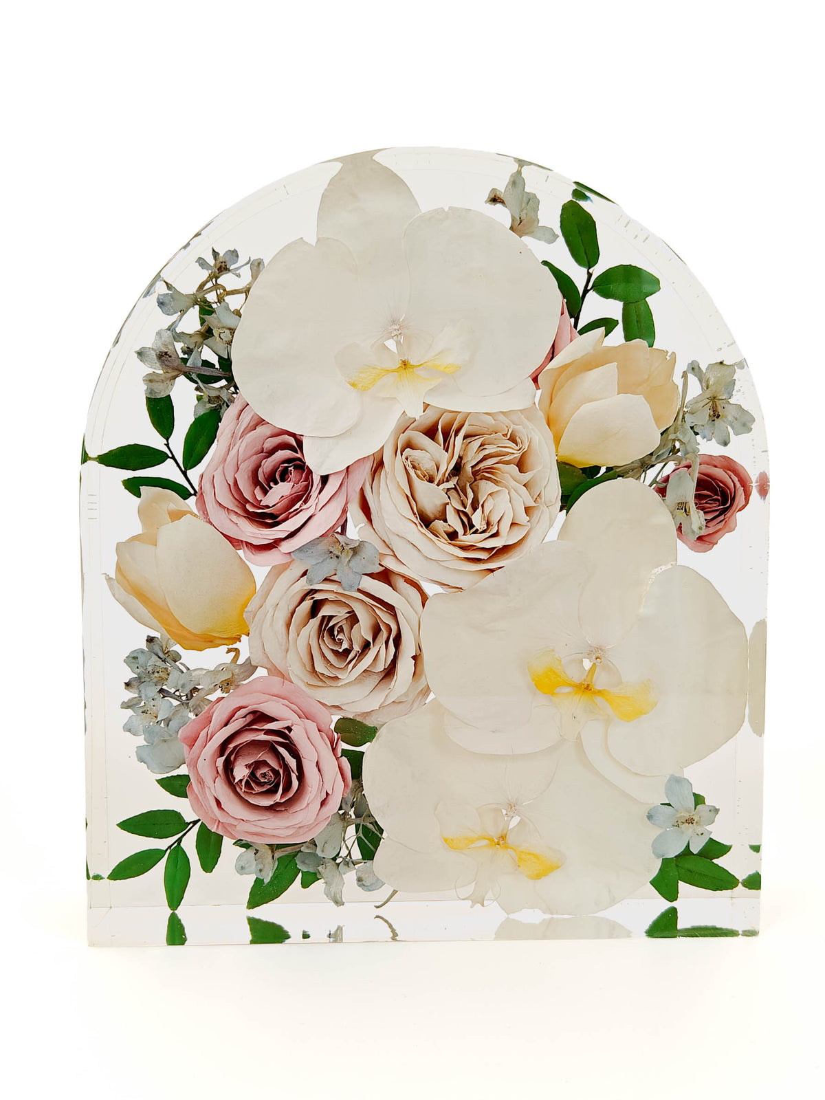 An arched clear resin plaque featuring a pressed floral arrangement with white orchids, pink and beige roses, green leaves, and small light blue flowers, photographed on a white background.