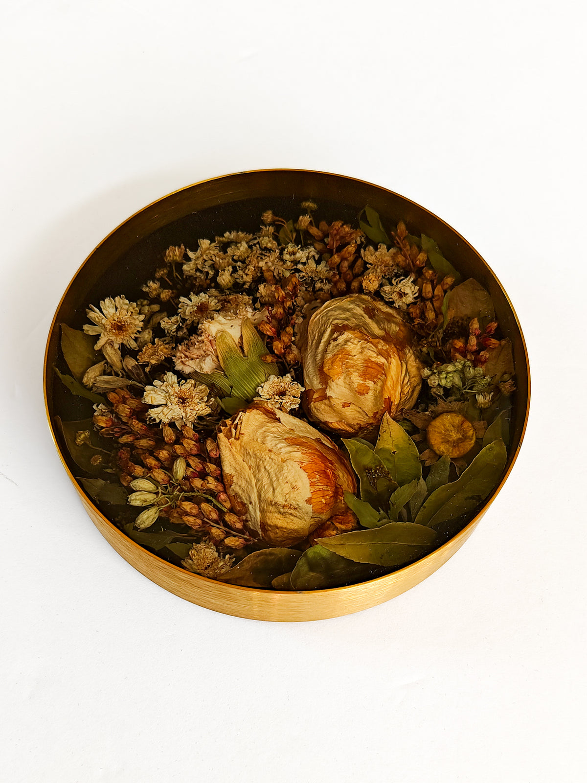 A round gold-toned metal tin filled with dried botanicals, including white flowers, green leaves, and tan seed pods, photographed from above on a plain white background.