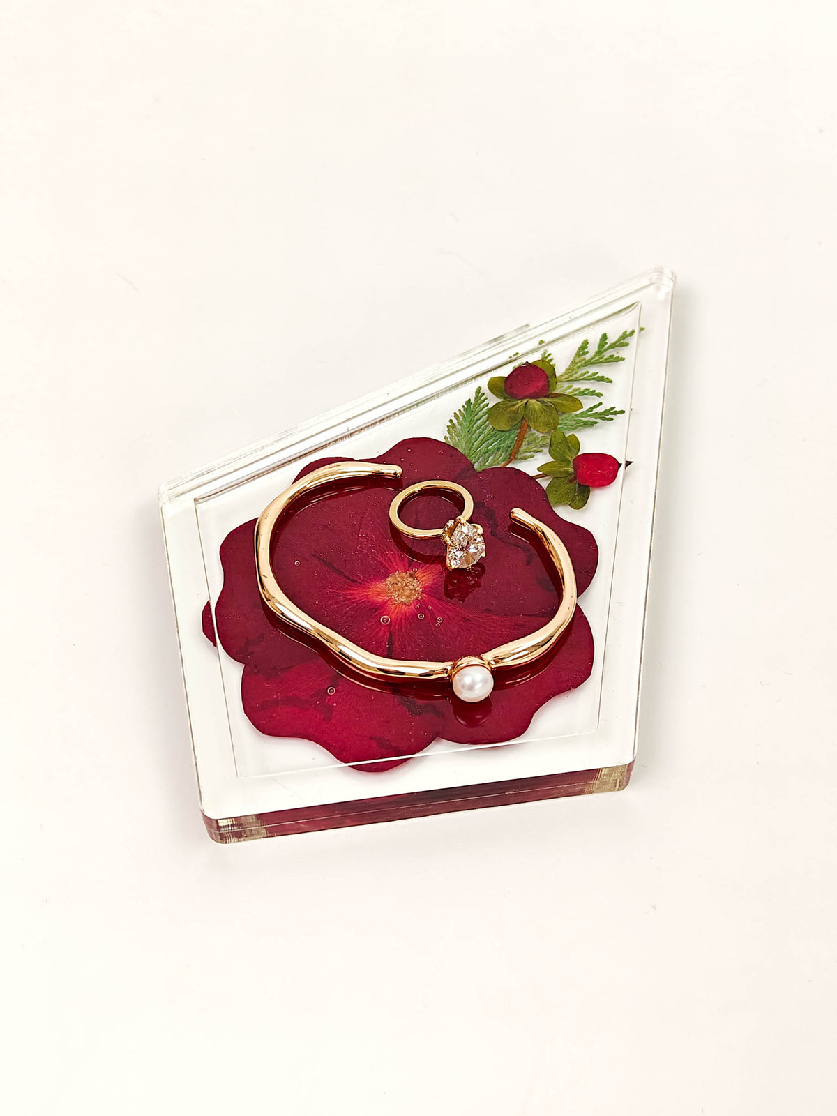A gold-toned open cuff bracelet with a small white pearl bead and a matching gold ring with a clear stone, displayed on a red flower-shaped insert inside a clear acrylic block against a white background.