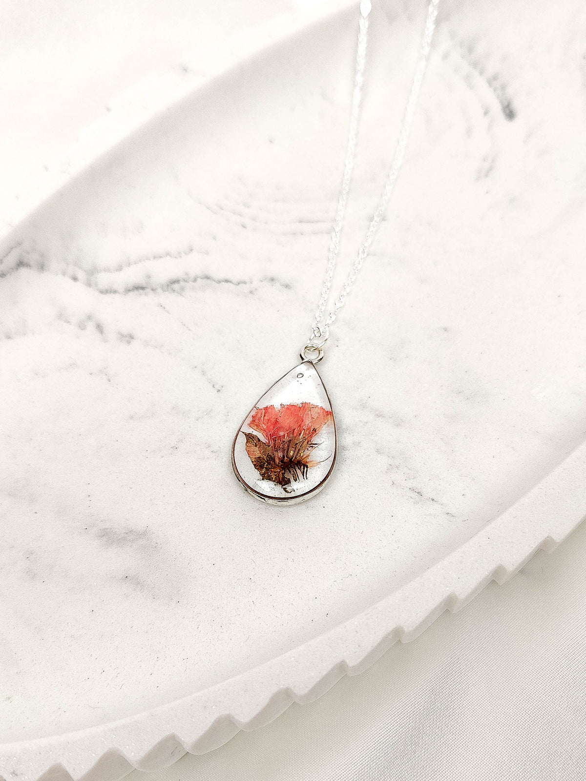 A teardrop-shaped pendant necklace on a delicate silver chain, featuring a dried orange and brown flower preserved inside a clear resin dome, displayed on a white marble tray.