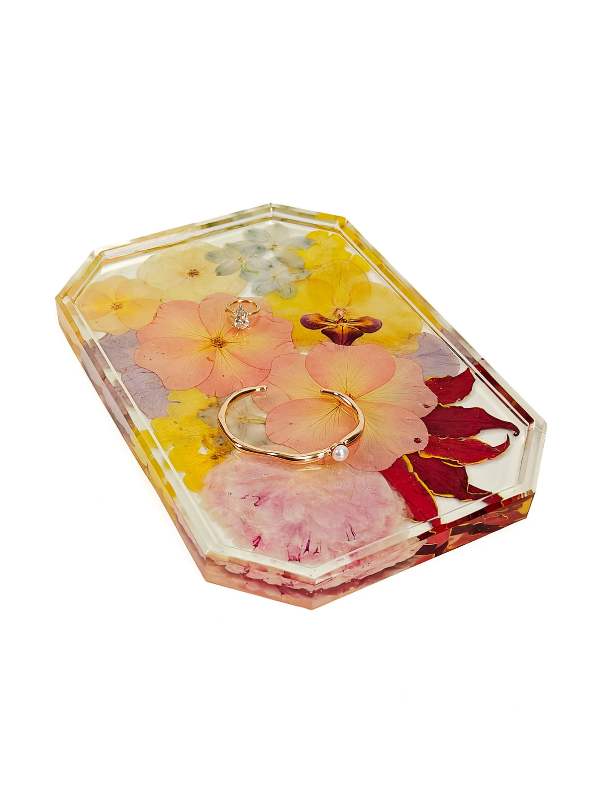 A clear acrylic keepsake box with a hinged lid, filled with pressed flowers in shades of pink, yellow, and red, and featuring a small gold ring with a pearl accent resting inside, set against a plain white background.