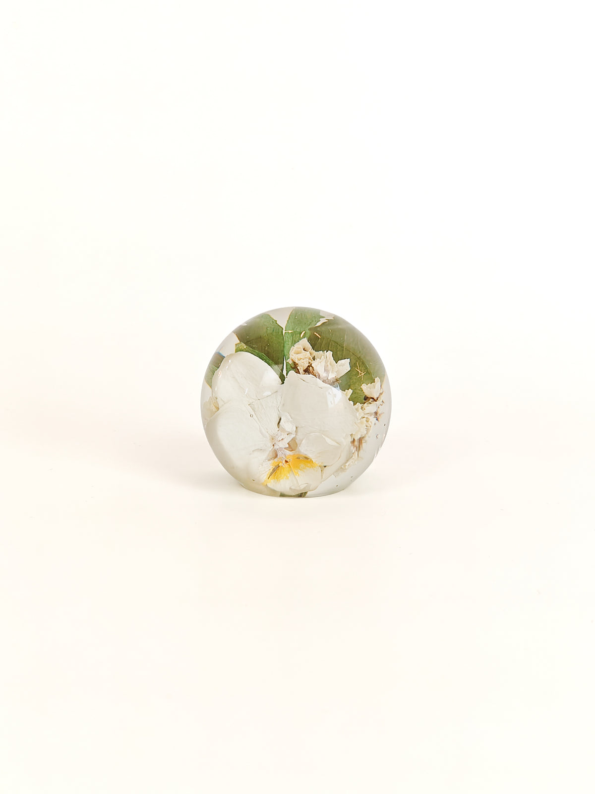 A small round glass paperweight featuring an embedded floral design with white blossoms, green leaves, and a touch of yellow, set against a plain white background.