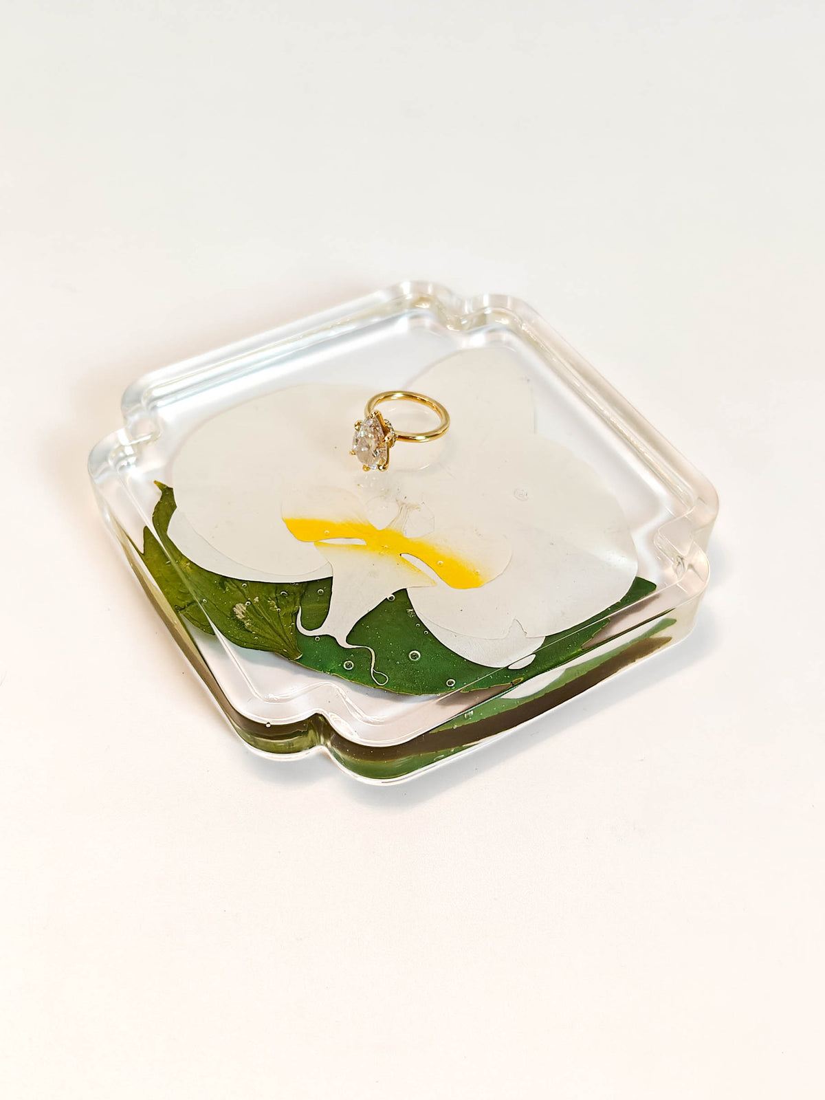 A clear acrylic ring holder shaped like a white flower with green leaves and a yellow center, containing a gold ring, photographed on a clean white background.