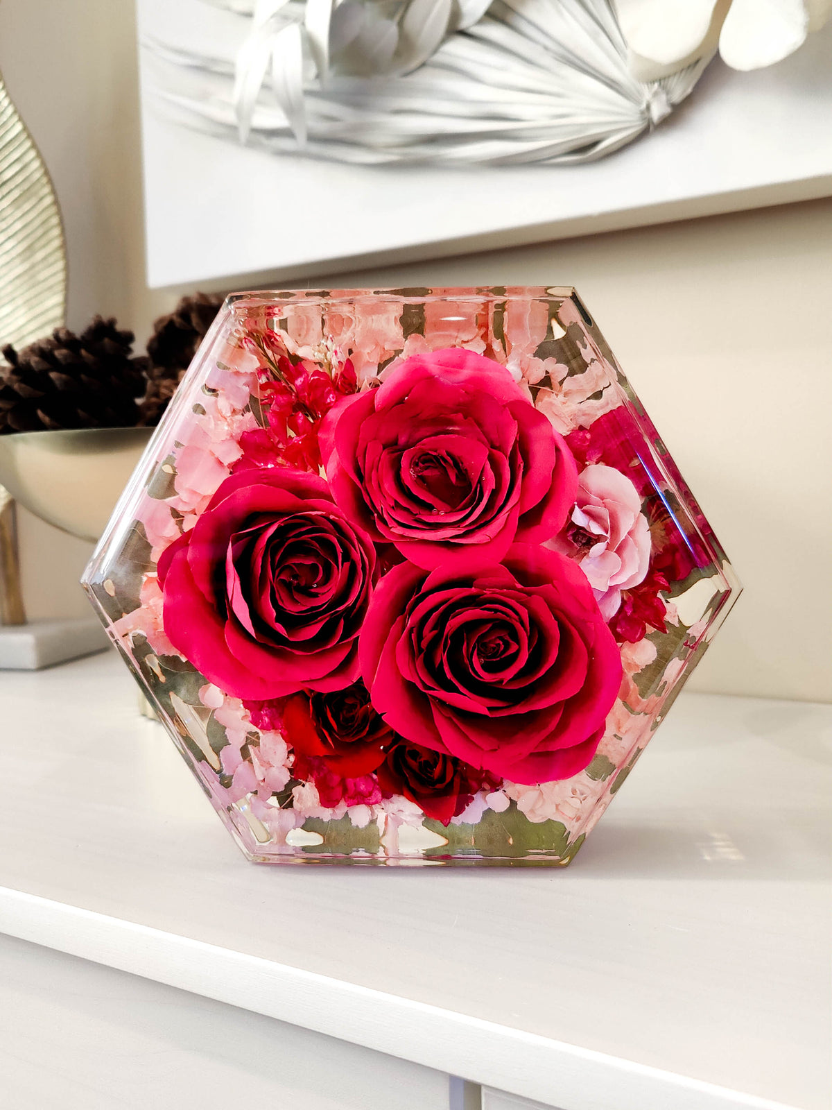 A hexagonal clear resin vase containing three large pink roses and small pink filler flowers, displayed on a white shelf in an indoor setting.