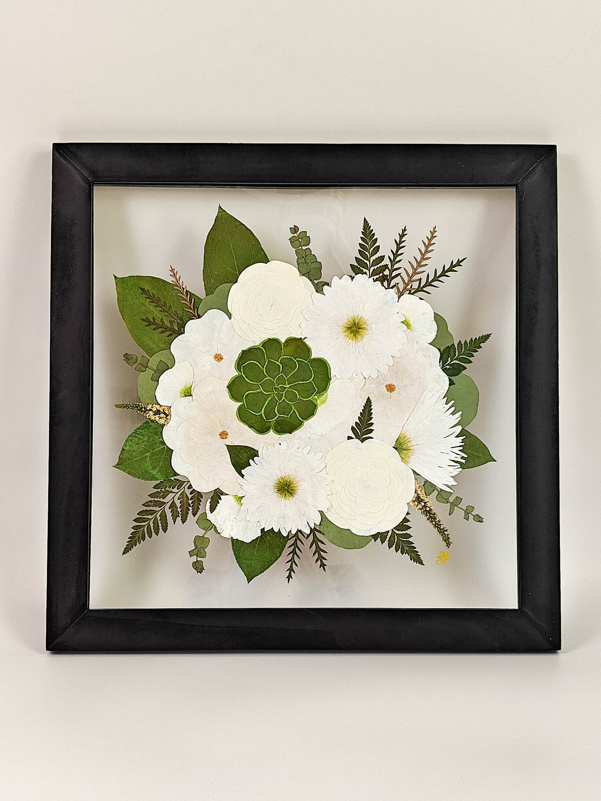 A framed pressed flower arrangement featuring white blossoms, green leaves, and delicate ferns inside a black square frame, displayed against a plain light beige wall.