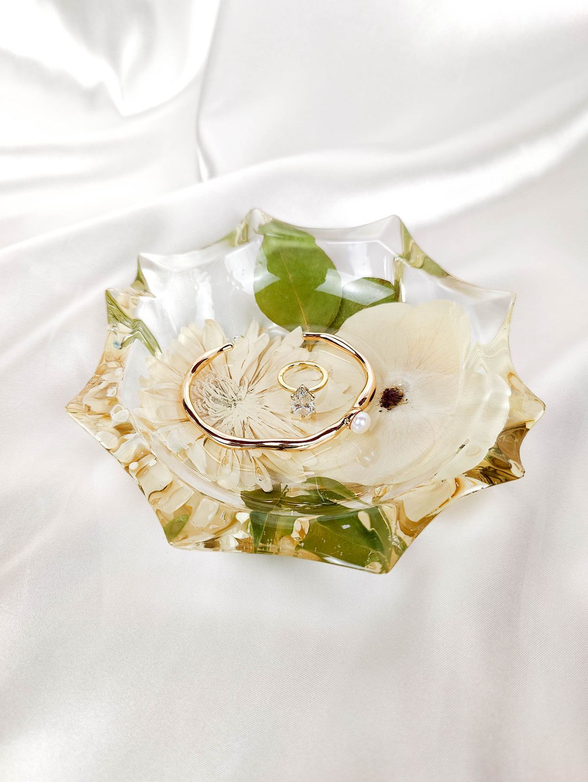 A clear resin jewelry tray with a scalloped edge, containing dried cream flowers and green leaves, styled with gold rings on a white satin fabric background.
