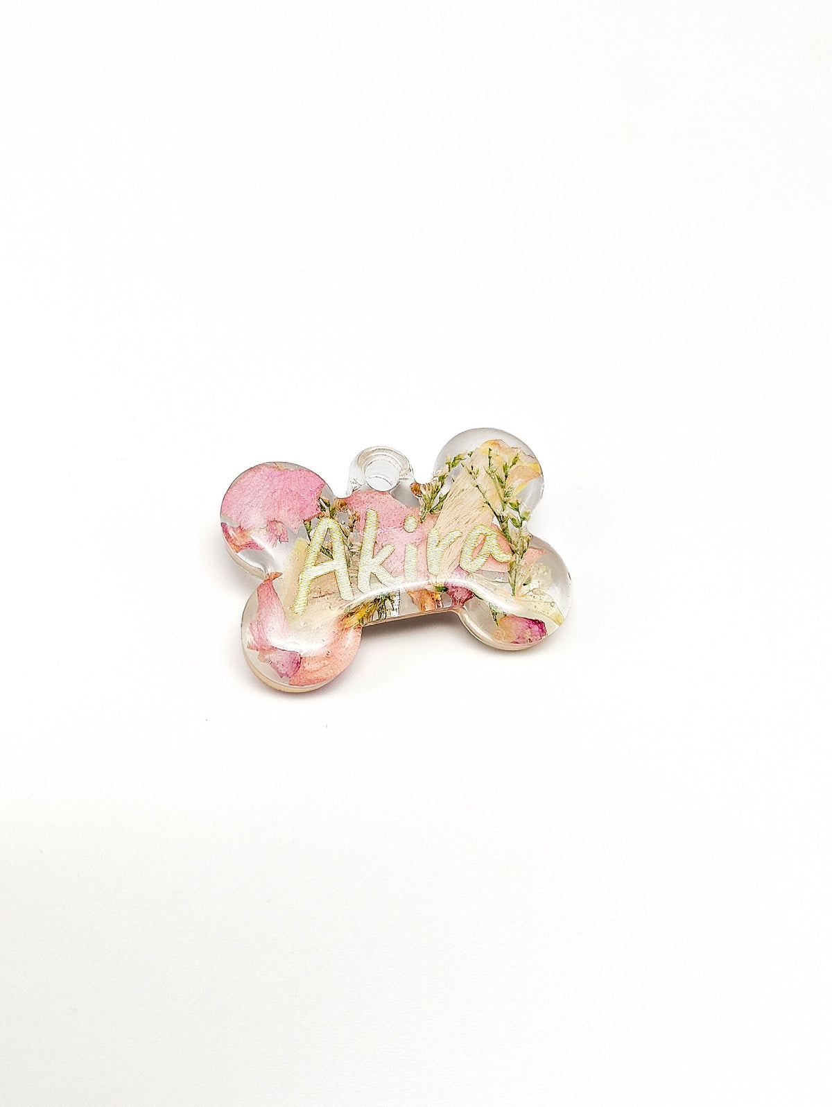 A bone-shaped pet ID tag with a pink and gold floral pattern, featuring the name 'Akila' in gold foil lettering, set against a plain white background.