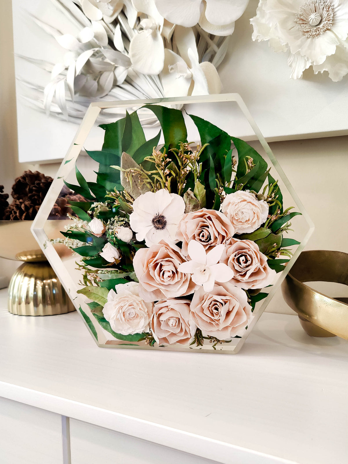 A hexagonal clear resin display box containing a floral arrangement with blush pink roses, green leaves, and white flowers, placed on a white tabletop in a styled interior setting.