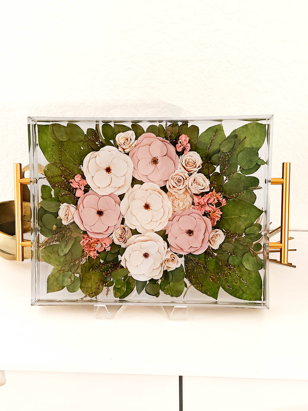 A rectangular clear acrylic shadow box tray with gold side handles, filled with a floral arrangement of pink and cream paper flowers and green leaves, displayed on a white surface.