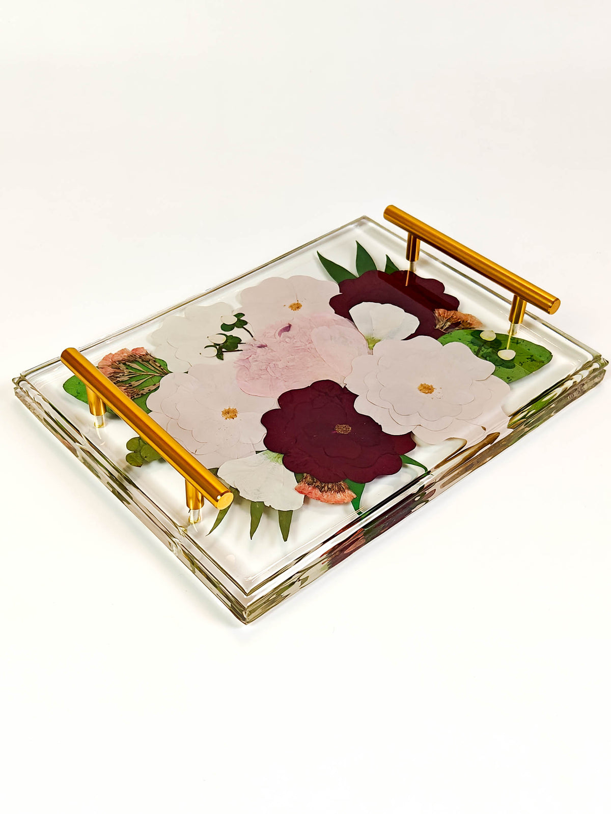 A rectangular clear glass serving tray with gold metal handles, decorated with pressed flowers in shades of white, burgundy, and pink with green leaves, set against a plain white background.
