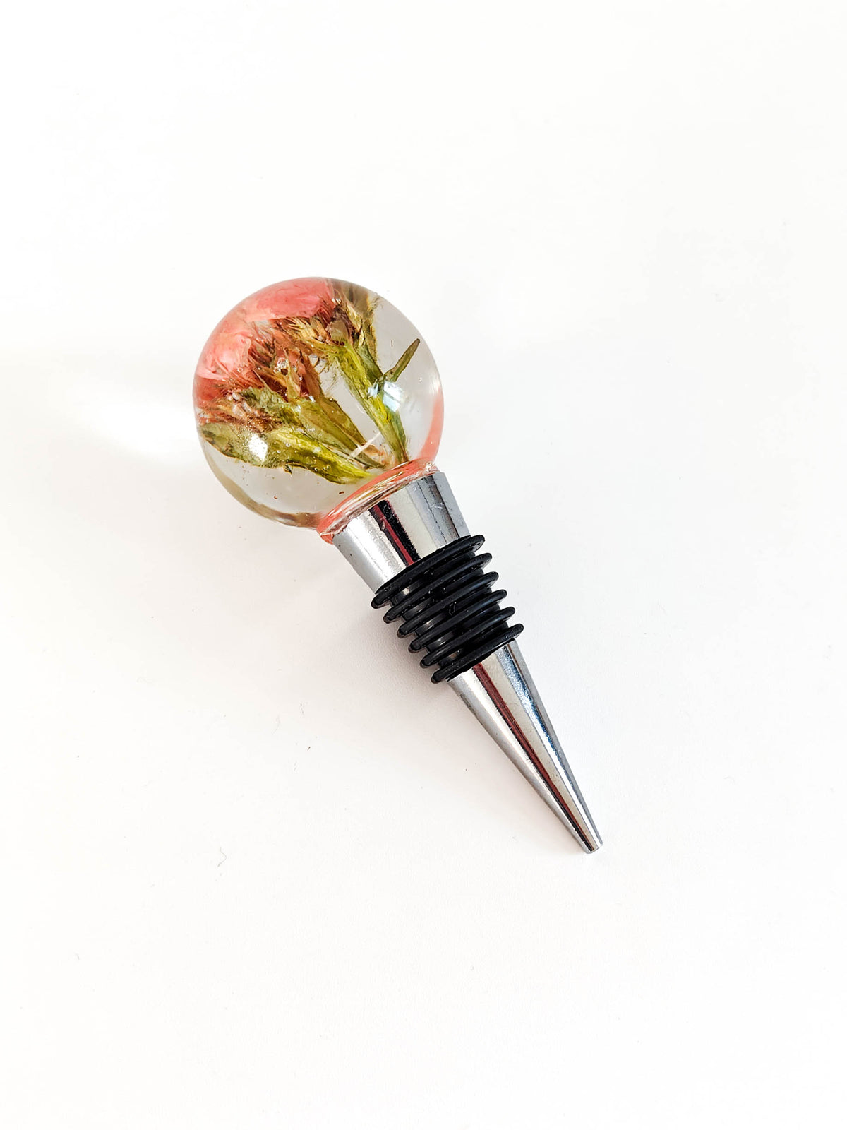 A bottle stopper featuring a clear spherical top containing dried pink and green botanical elements, connected to a silver metal cone-shaped point with a black ribbed rubber seal, shown on a plain white background.