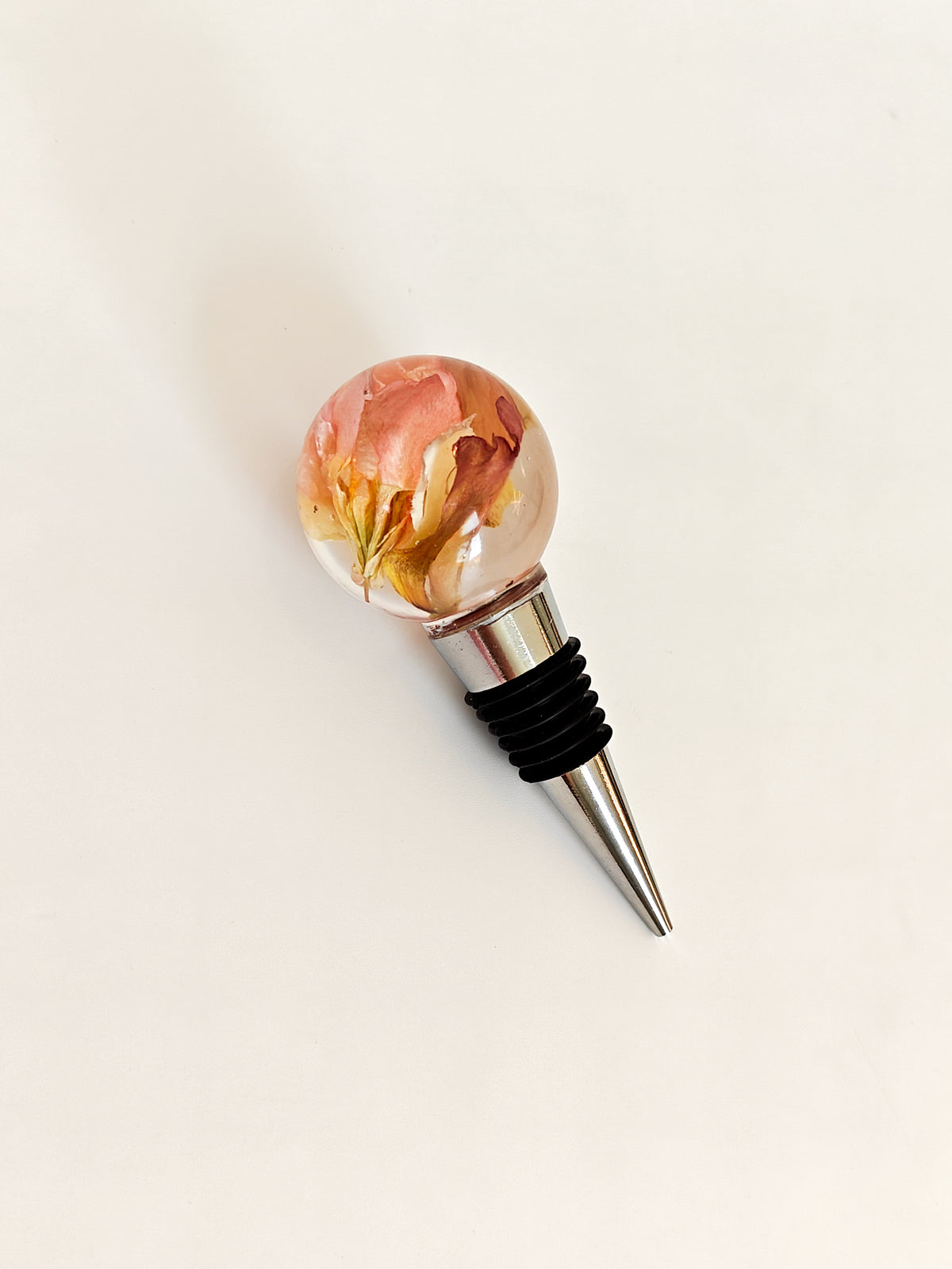 A wine bottle stopper featuring a clear glass sphere filled with dried pink and yellow flowers, attached to a silver metal spike with a black rubber gasket, shown on a plain off-white background.