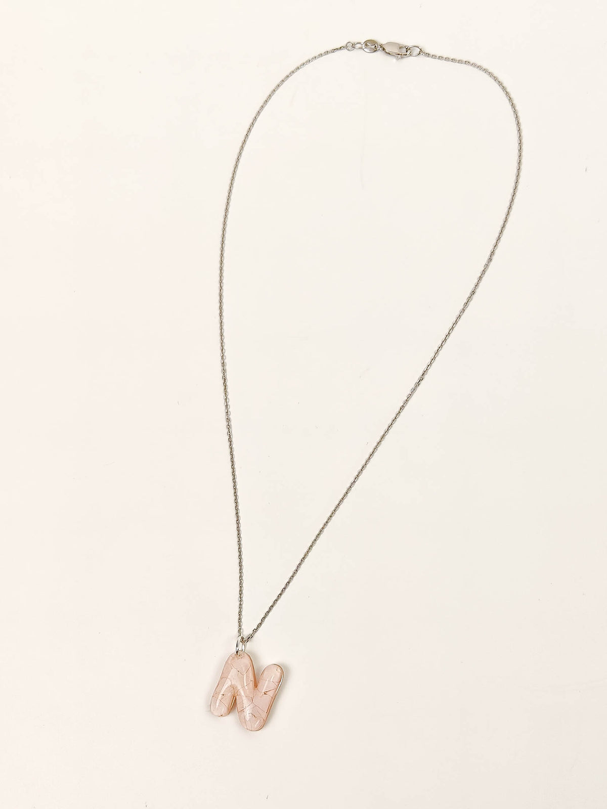 A delicate silver chain necklace featuring a small, polished peach-pink stone pendant shaped like the letter 'N', displayed on a clean, off-white background.