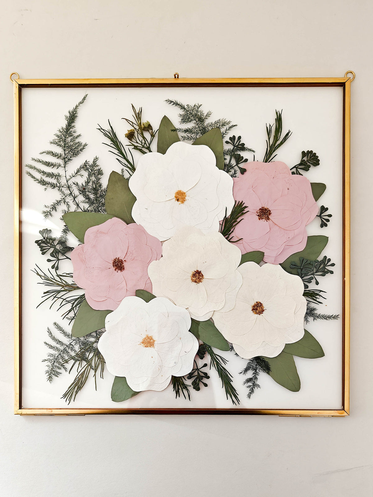 A framed floral arrangement featuring layered white and pink paper flowers with green leaves and pine branches, set in a gold frame against a light wall.