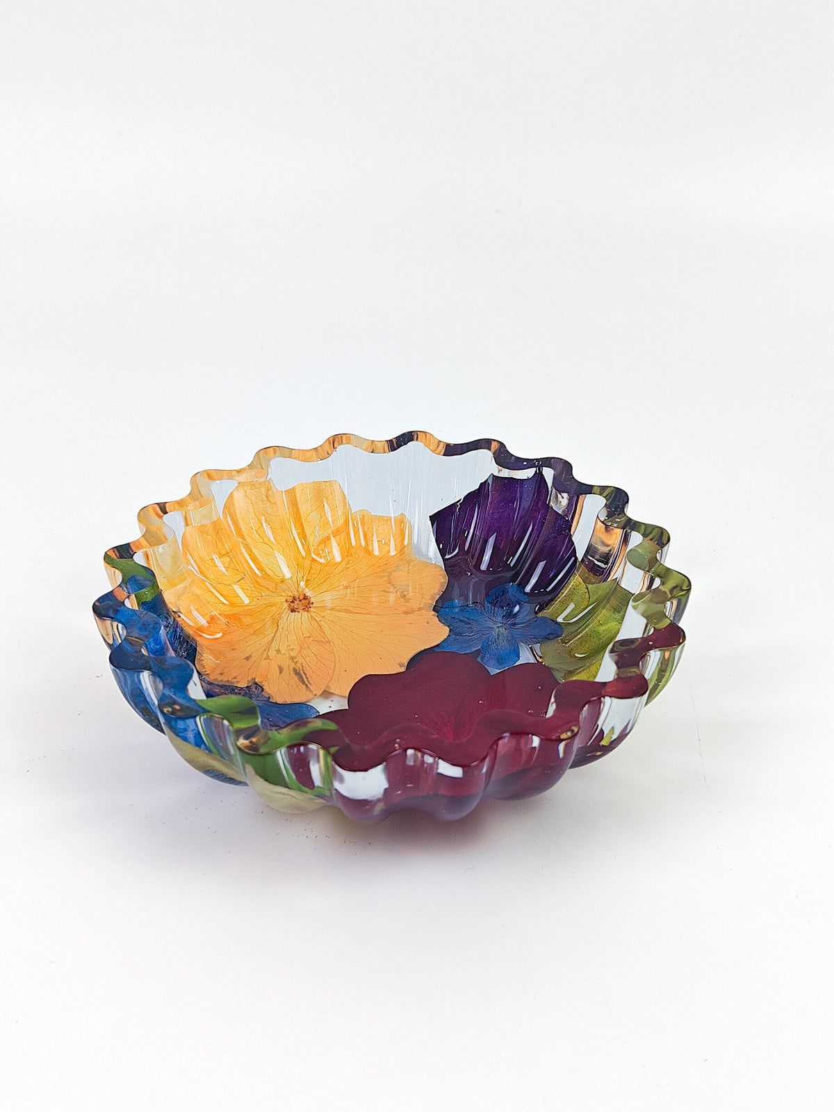 A scalloped resin bowl with a multicolor floral pattern, featuring yellow, purple, blue, green, and red petal-like sections, set against a plain light gray background.