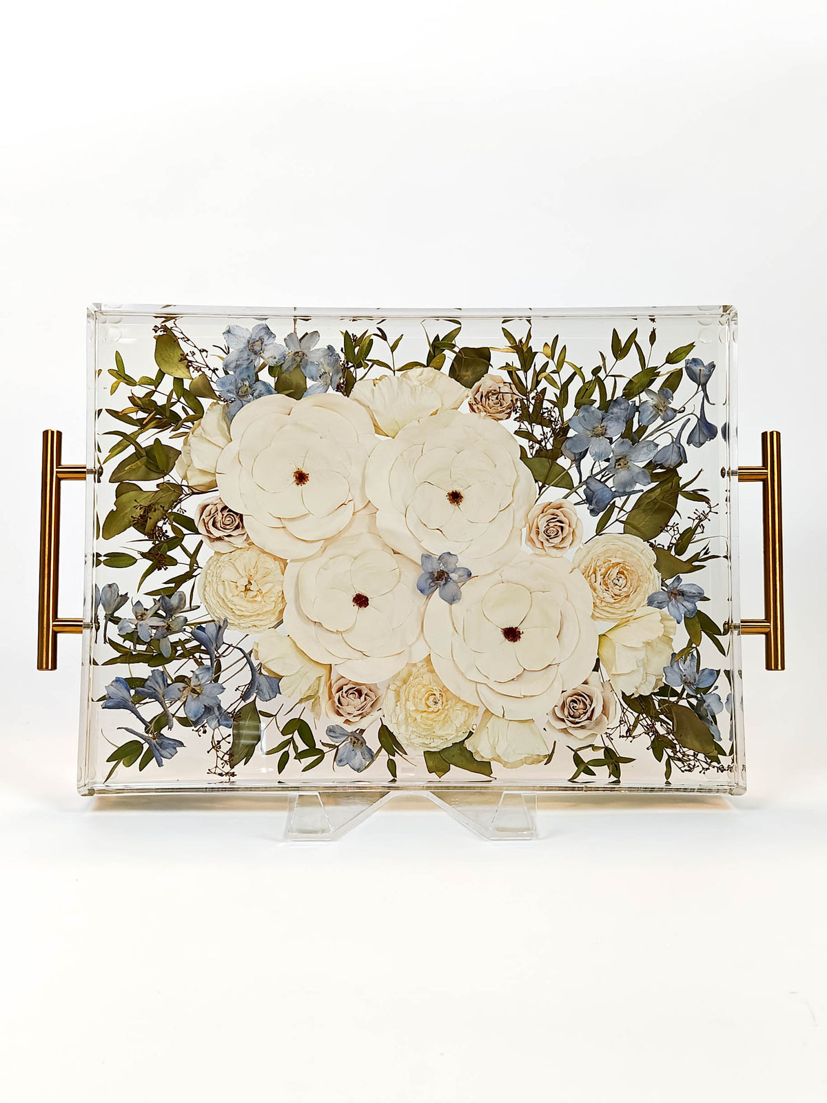 A rectangular clear acrylic decorative tray with gold-toned side handles, displaying a pressed floral arrangement of cream roses, pale yellow blooms, green leaves, and small blue flowers on a white background.