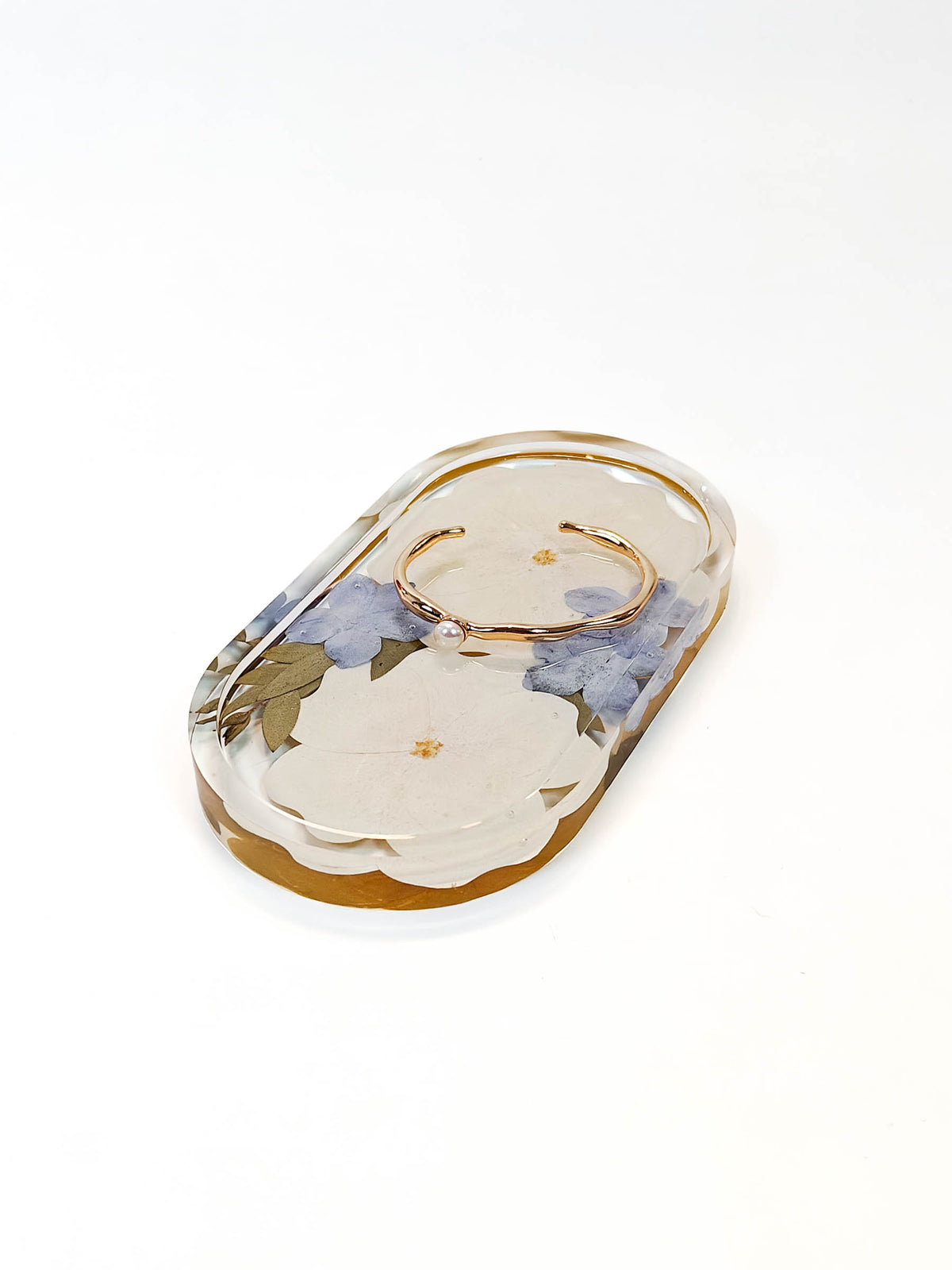 A clear resin ring holder tray with a scalloped edge, featuring dried white flowers, blue hydrangeas, and small gold foil flecks, holding a gold ring on a white background.