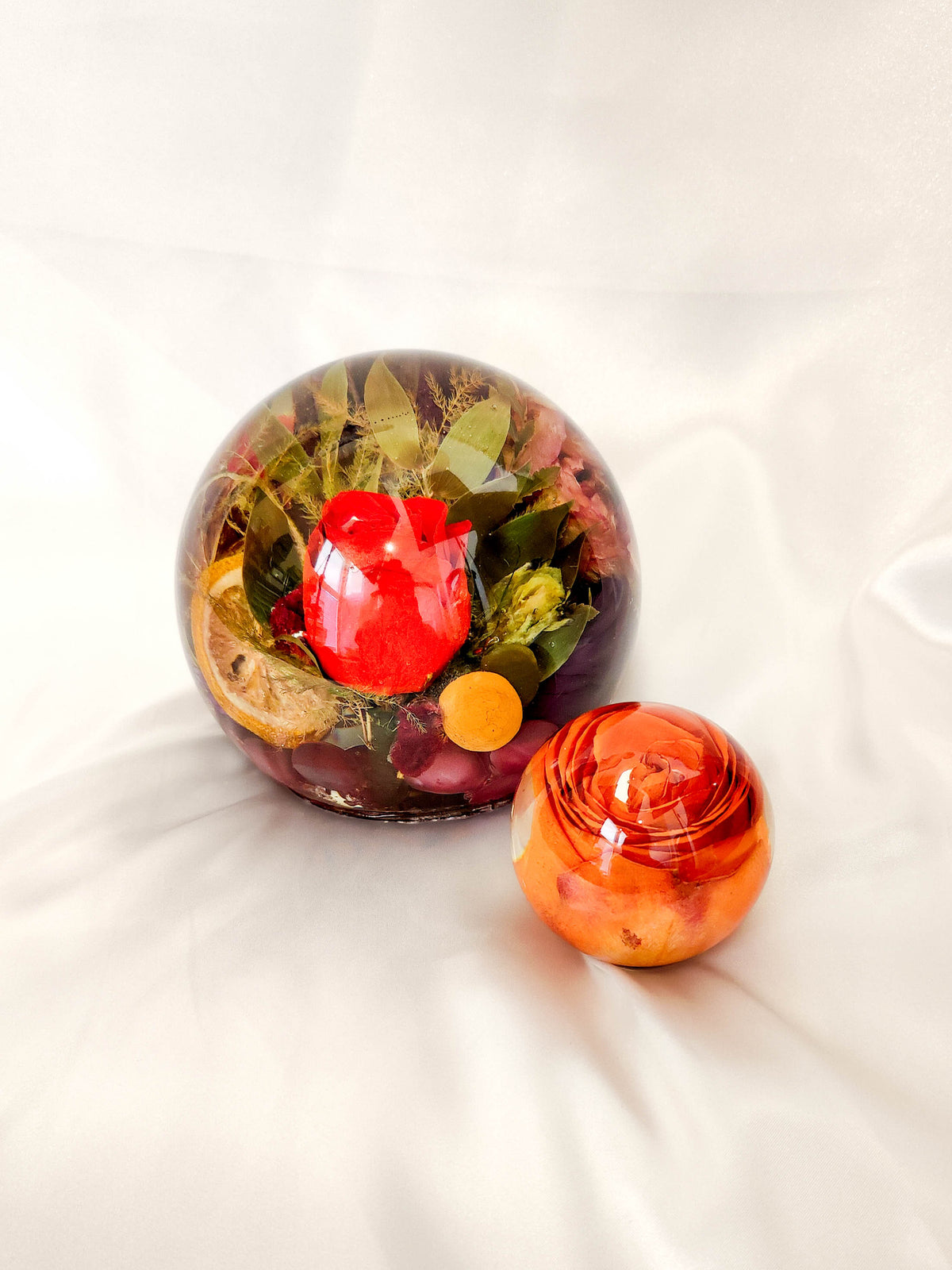 A decorative glass orb containing dried botanicals, leaves, and a bright red rose, shown next to a smaller orange marbled glass rose on a white fabric background.