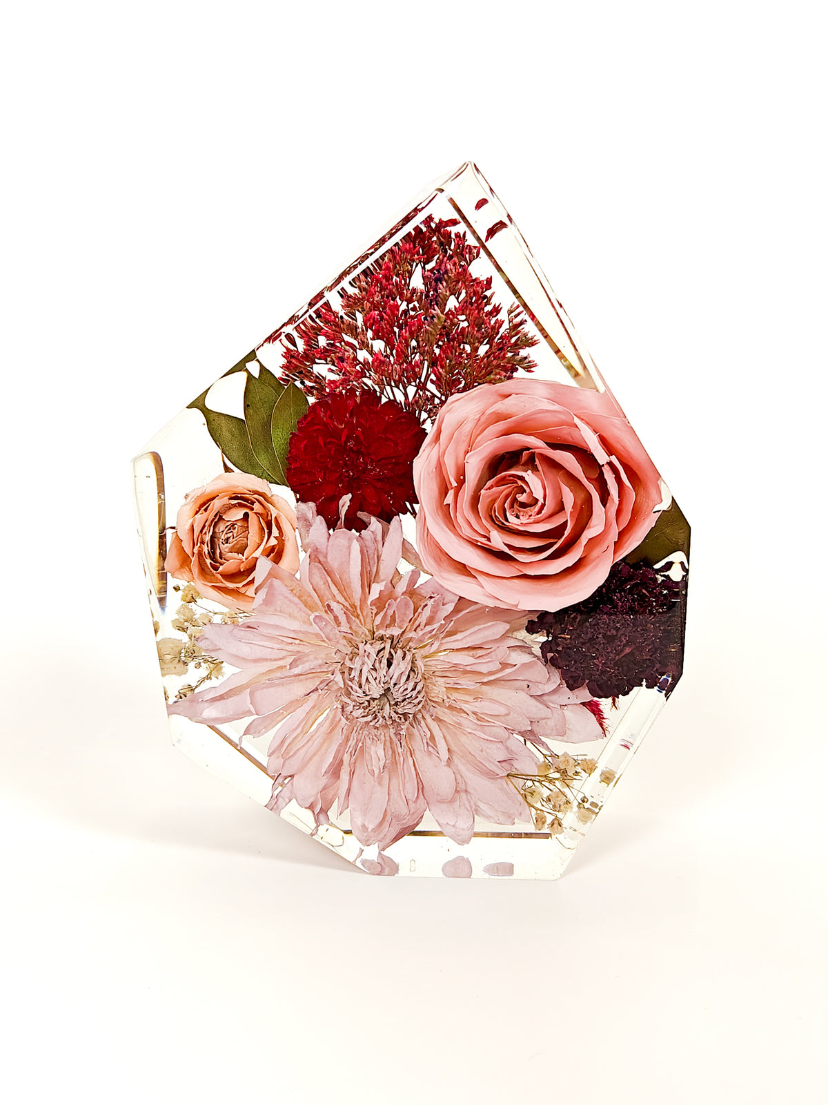 A clear resin geometric block with dried flowers, featuring a large pale pink dahlia, a pink rose, a peach rose, and red filler flowers, photographed on a white background.