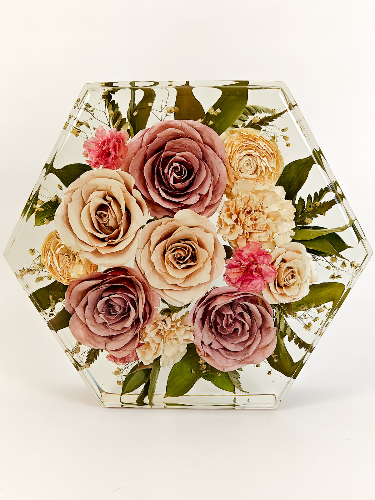 Hexagonal clear resin vase filled with a dried floral arrangement featuring mauve, cream, and pink roses, beige carnations, and green leaves, photographed on a white background.
