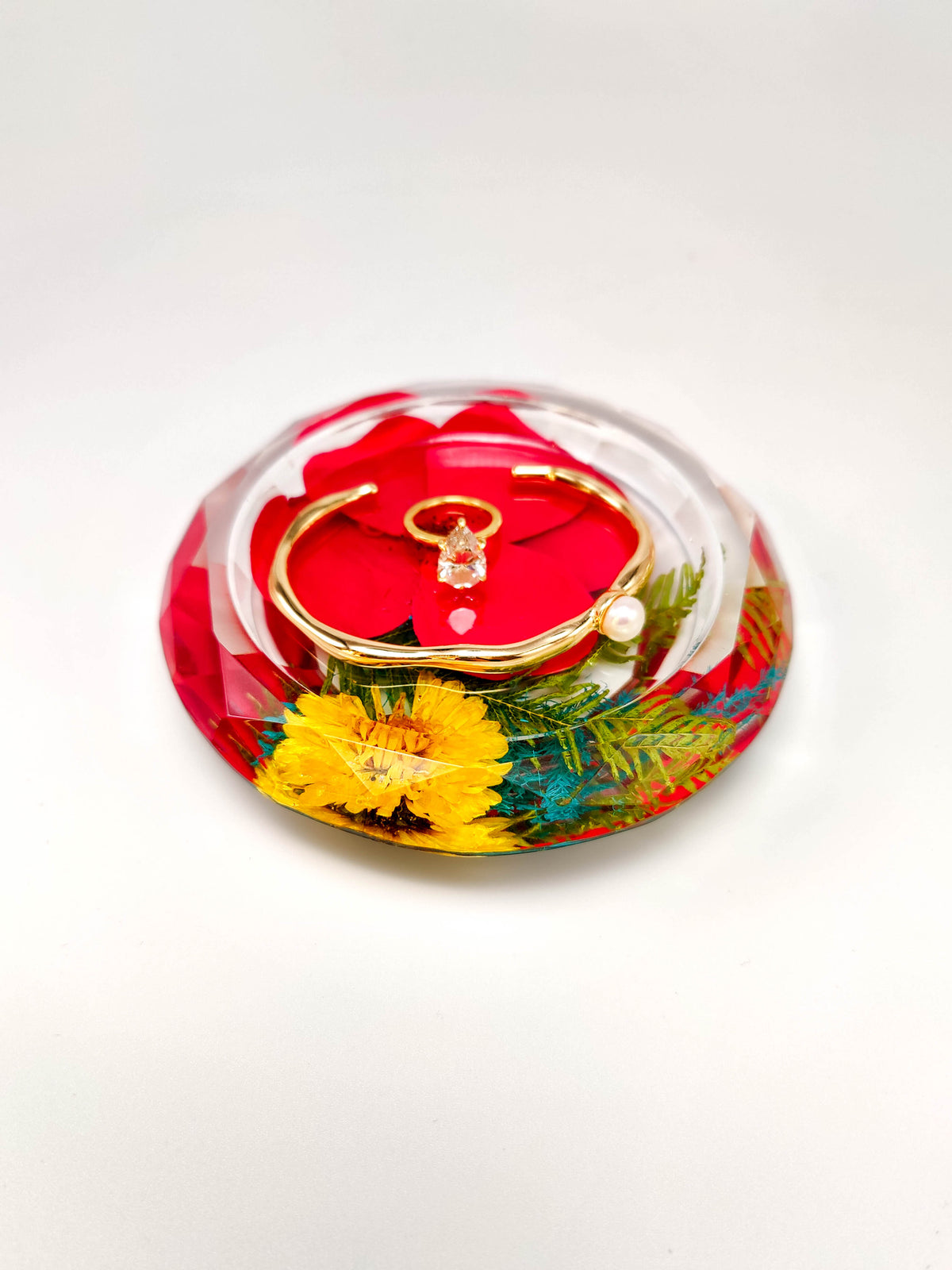 A gold-tone cuff bracelet with a clear crystal pendant and a small pearl accent, displayed inside a circular clear resin holder filled with red flowers and yellow botanical details, on a white background.