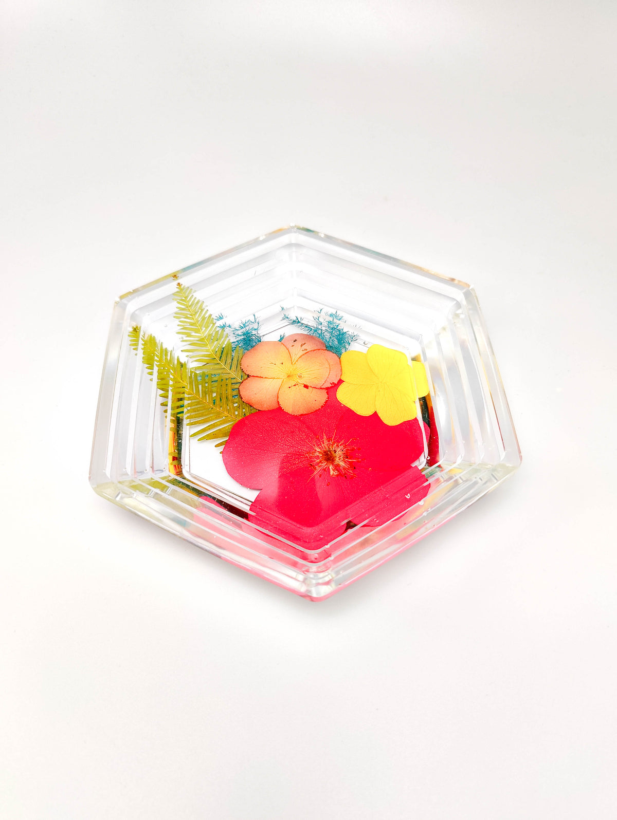 A clear hexagonal glass dish with a ribbed edge, containing pressed flowers and green leaves, photographed on a clean white background.