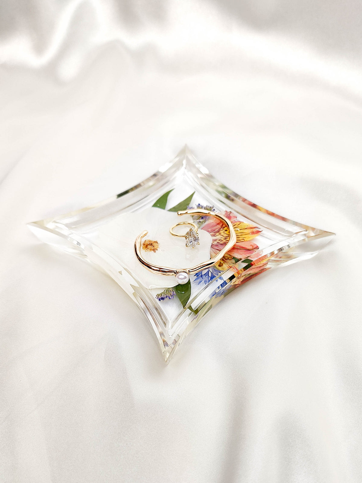 A clear acrylic ring box with a diamond-shaped top, decorated with pressed flowers and green leaves, holding a gold ring on a white satin background.