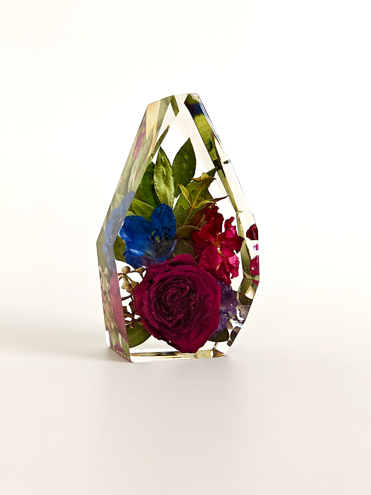 A clear faceted crystal or resin vase containing preserved dried flowers, including a prominent deep magenta rose, blue flowers, and green leaves, set against a plain white background.