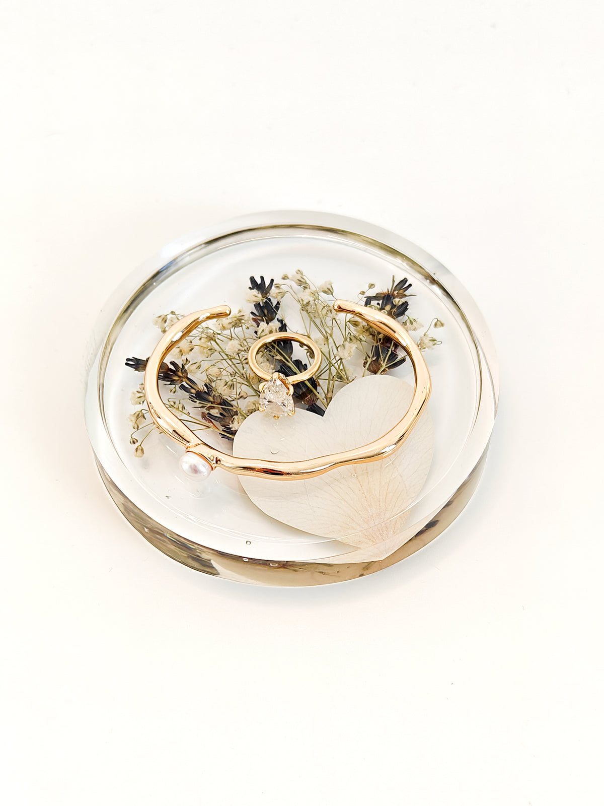 A gold-toned bangle bracelet with a small pearl accent and a matching ring, displayed inside a clear oval resin holder with dried flowers on a white background.
