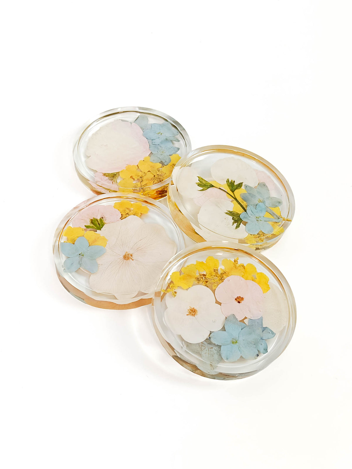 Four round clear resin coasters with embedded pressed flowers in white, yellow, and light blue, arranged on a white background.
