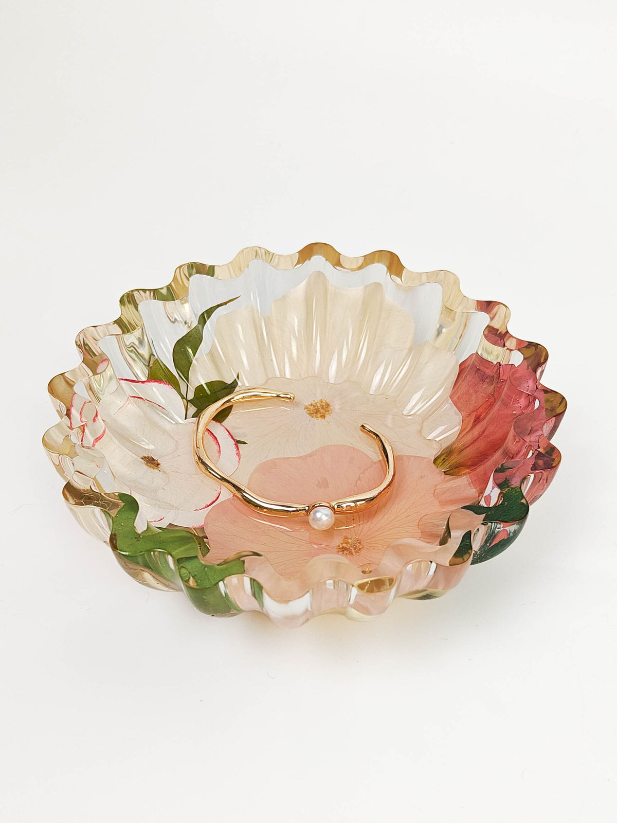 A gold open cuff bracelet with a small pearl accent resting inside a scalloped, floral-patterned glass bowl on a white background.